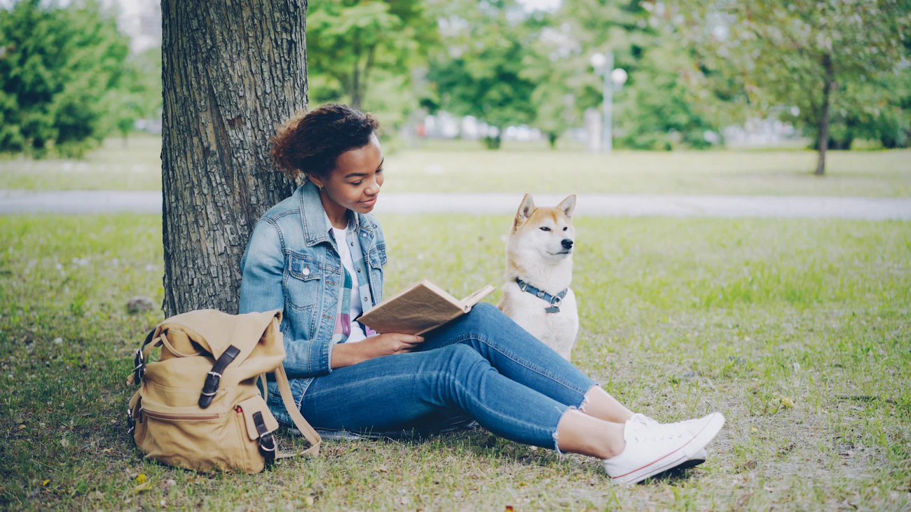 A young student with a backpack reading a book under a tree on a green campus lawn while her dog sits beside her