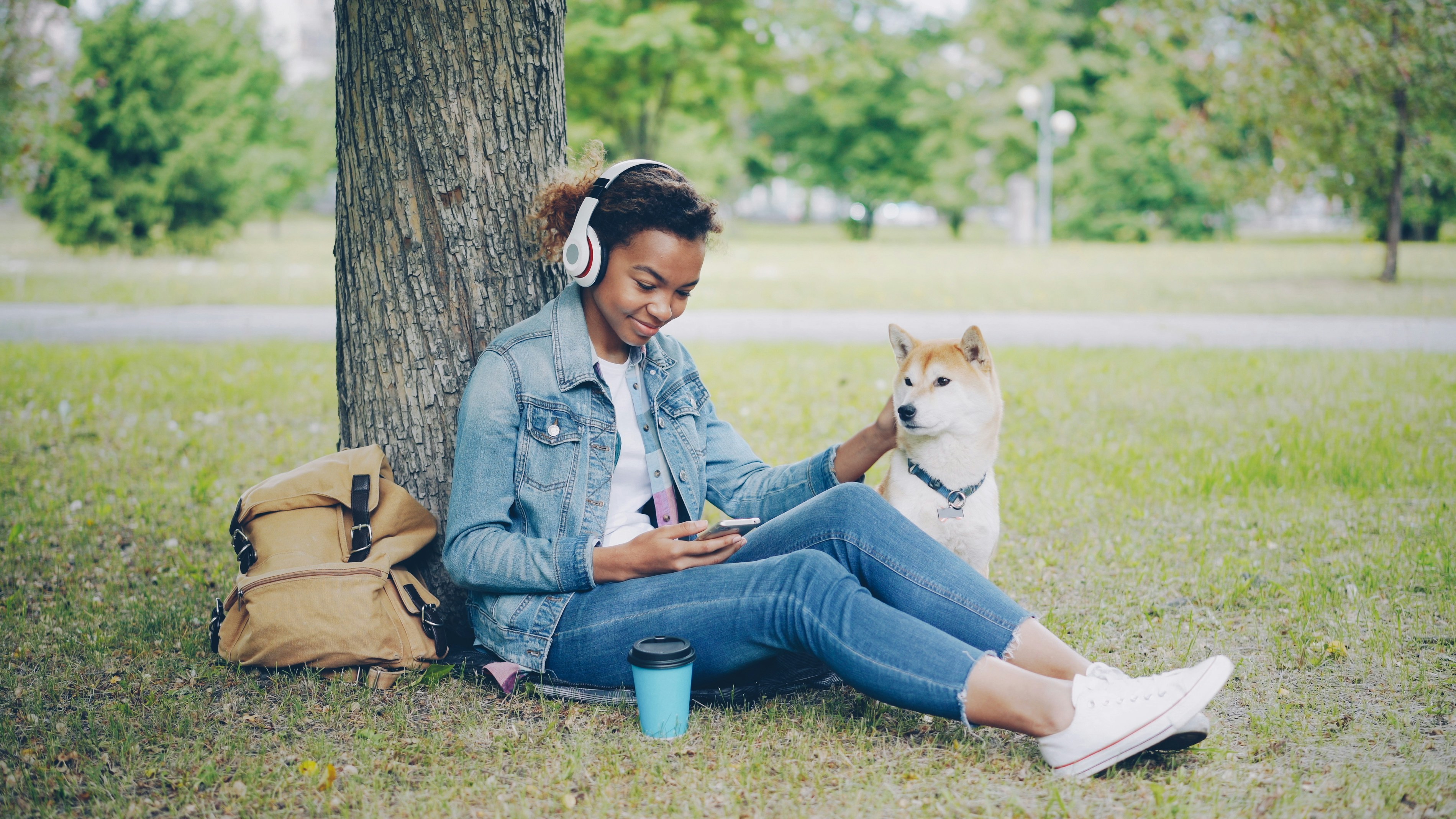 A young woman sits on the lush green grass, leaning comfortably against a large tree trunk in a park