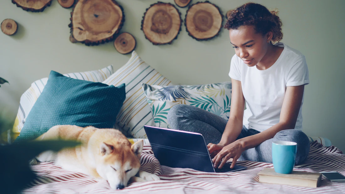 A student sitting on her residence bed with a laptop while her dog rests beside her