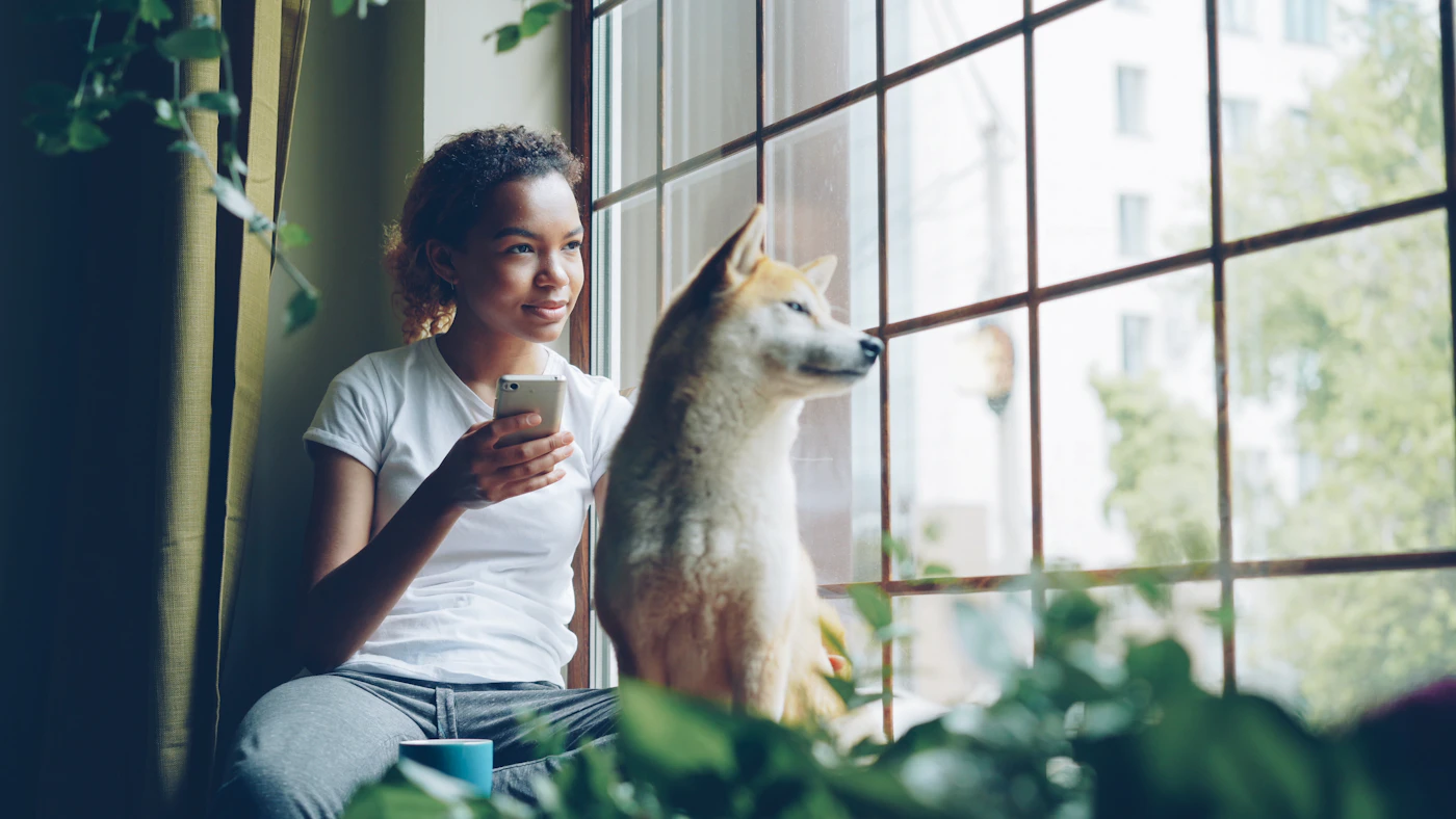 A woman and her dog resting at a window together, looking out at the quiet light of the morning