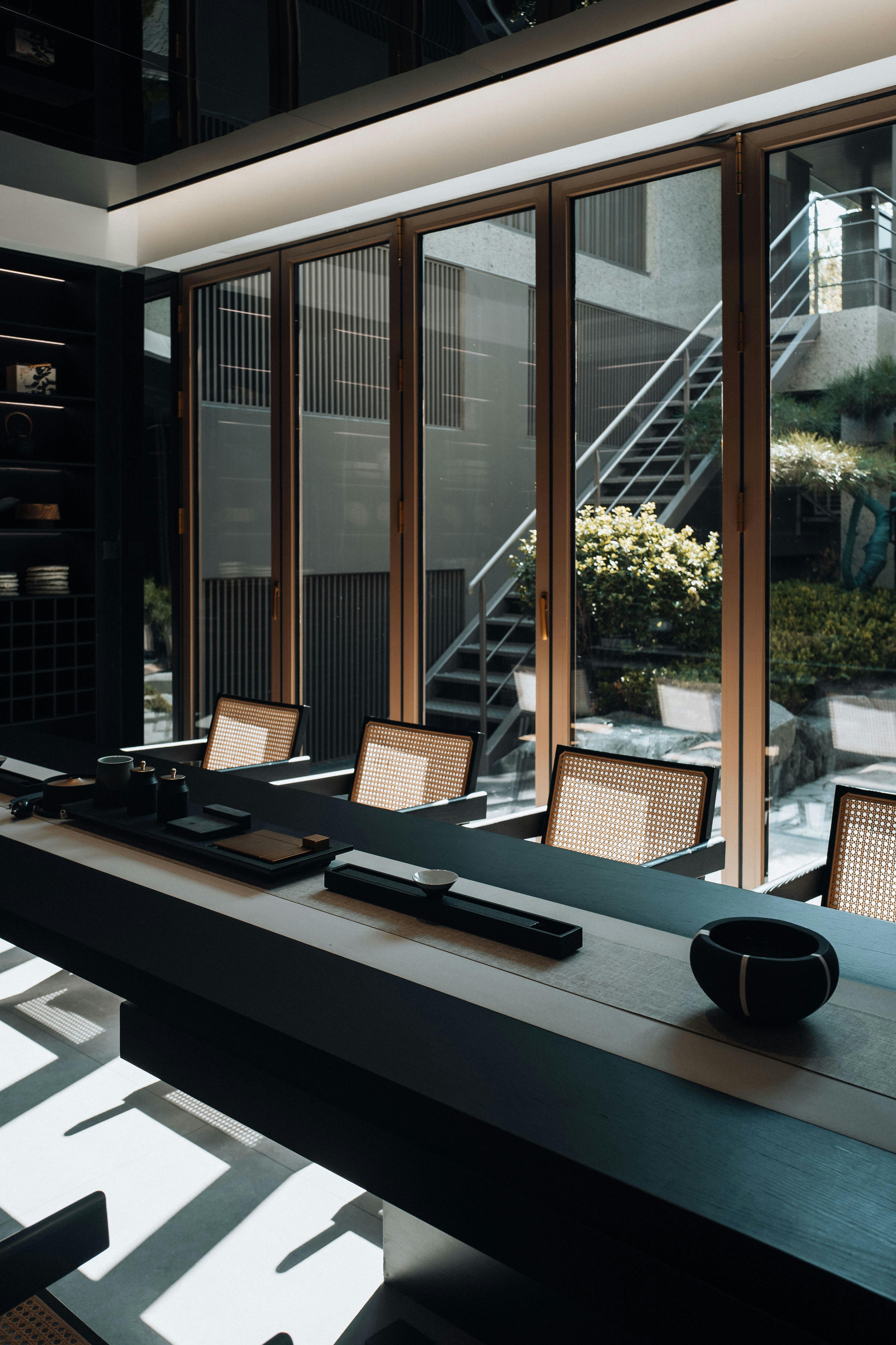 A long, dark wooden table with several light-colored chairs sits in a modern room with large glass doors that look out onto an outdoor area with stairs and greenery.