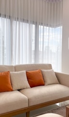 A couch with orange pillows sits by a window.