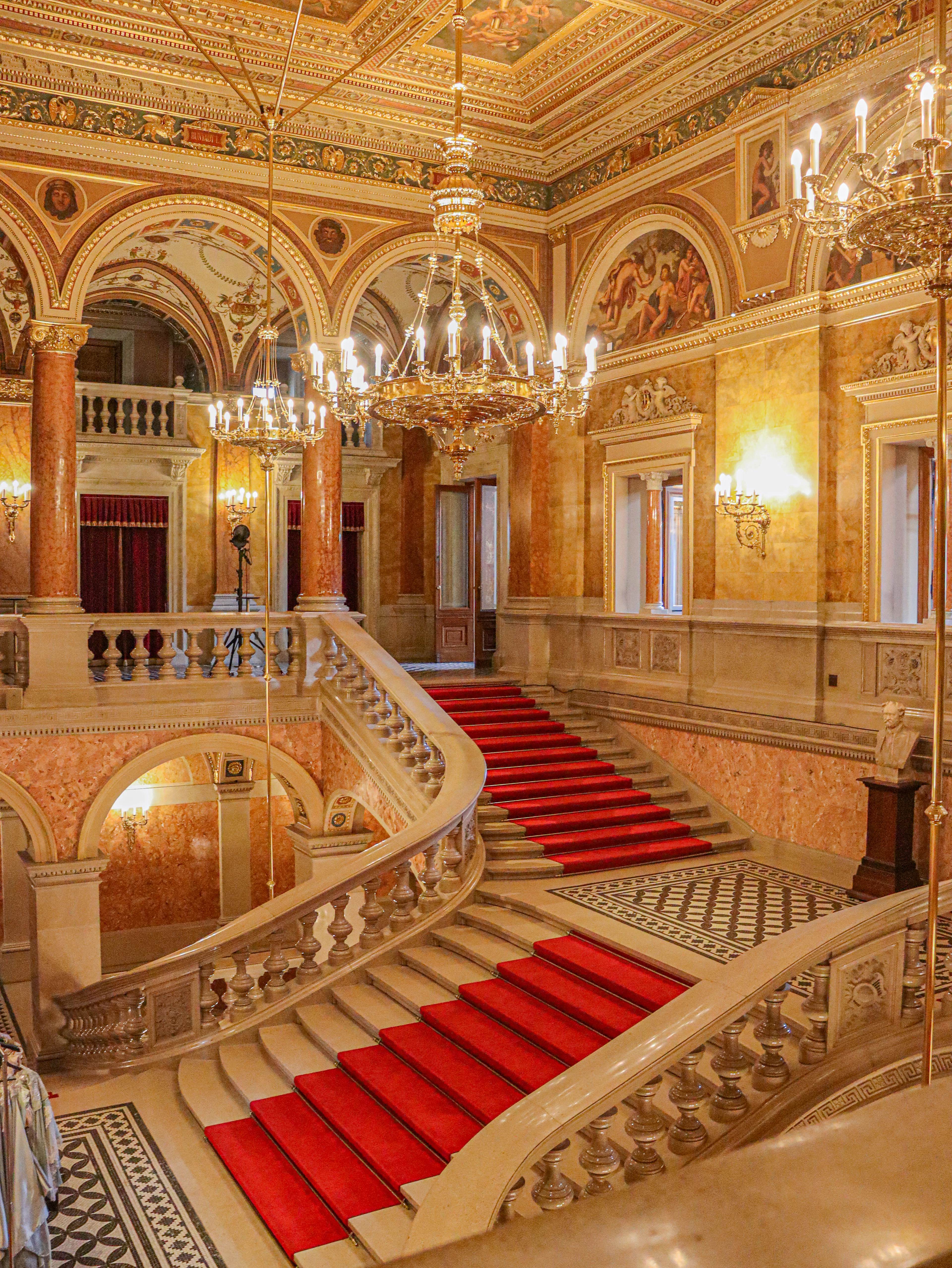 The grand staircase of the Hungarian State Opera House in Budapest, red carpet climbing between pale stone balustrades beneath frescoed vaults, marble columns and lit crystal chandeliers