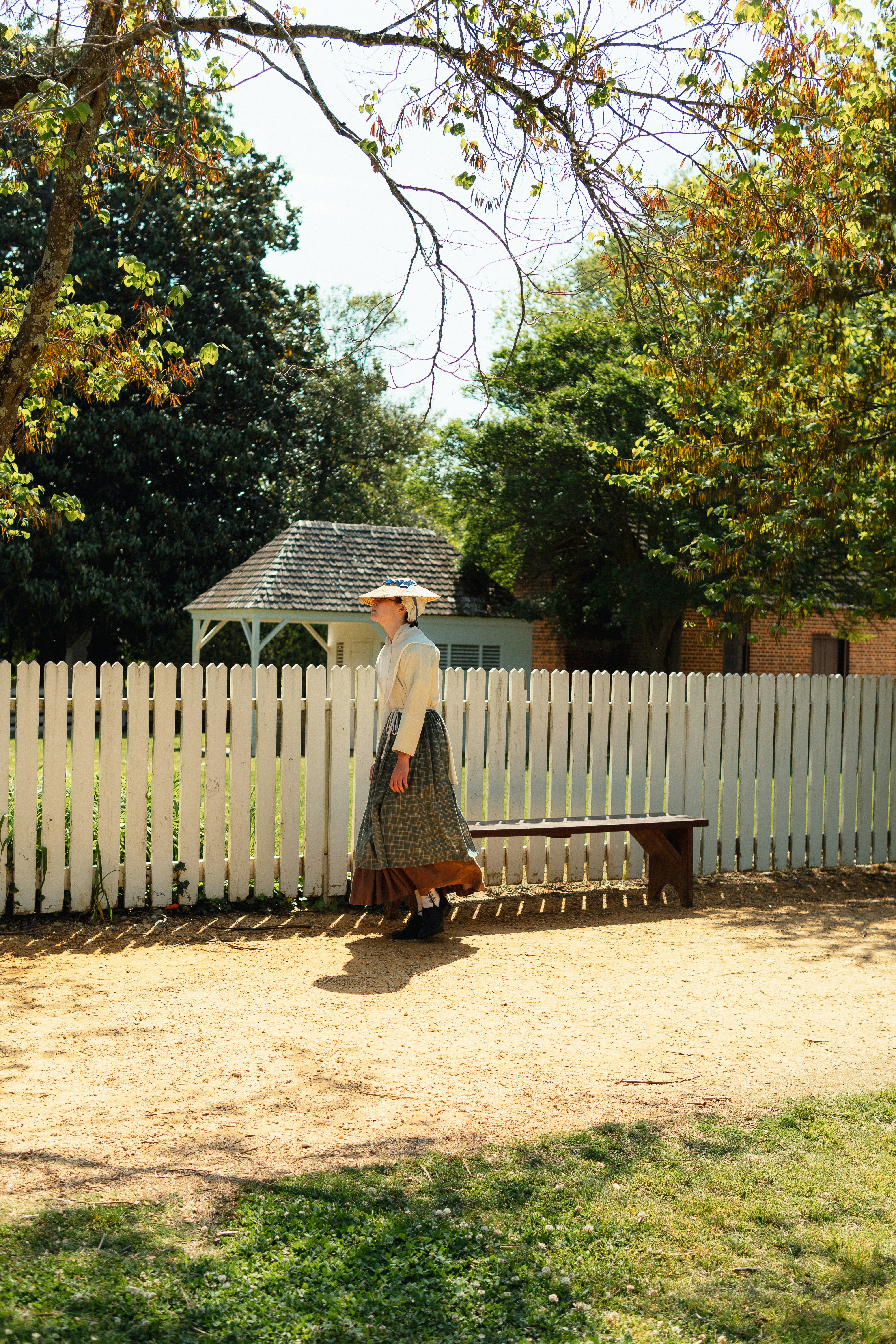 A woman in old attire walks near a white fence.