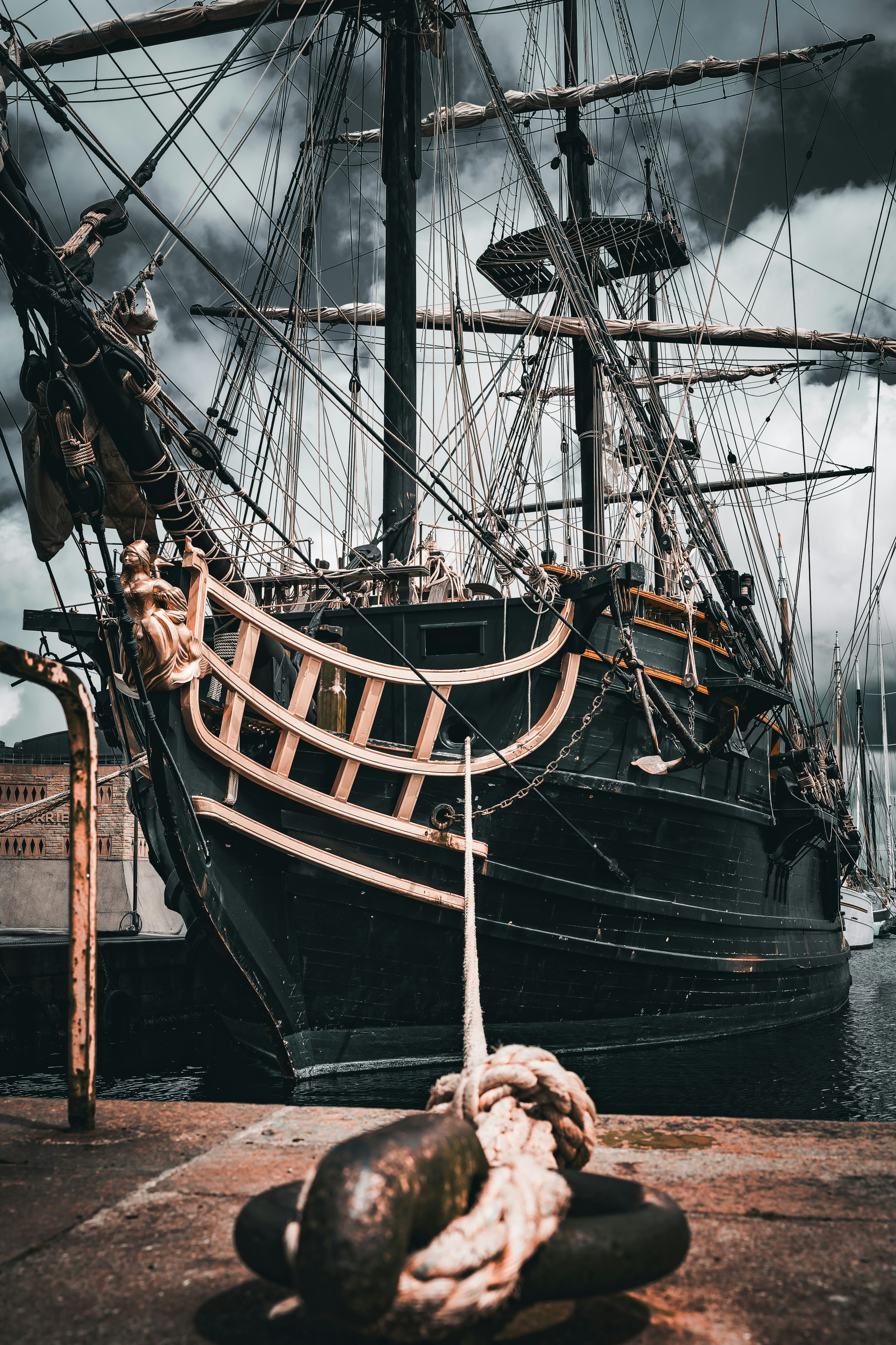 A tall ship sits docked under ominous, dark clouds.
