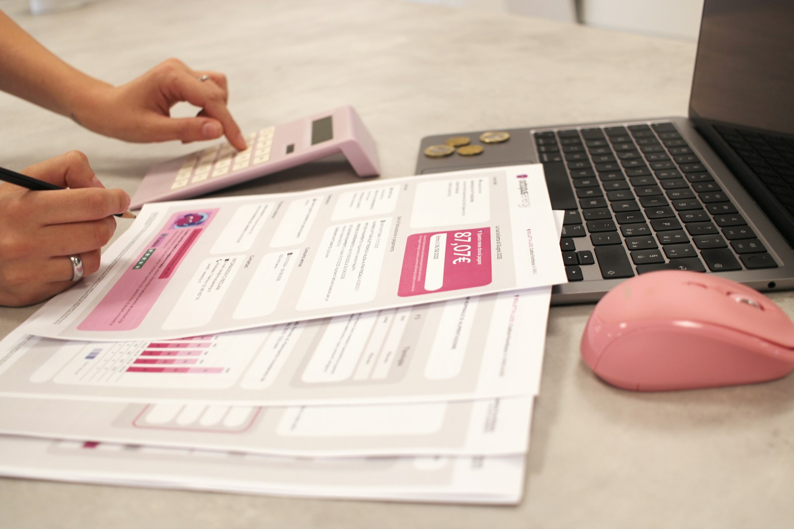 A person calculating bills beside a laptop, documents, coins, and a pink mouse