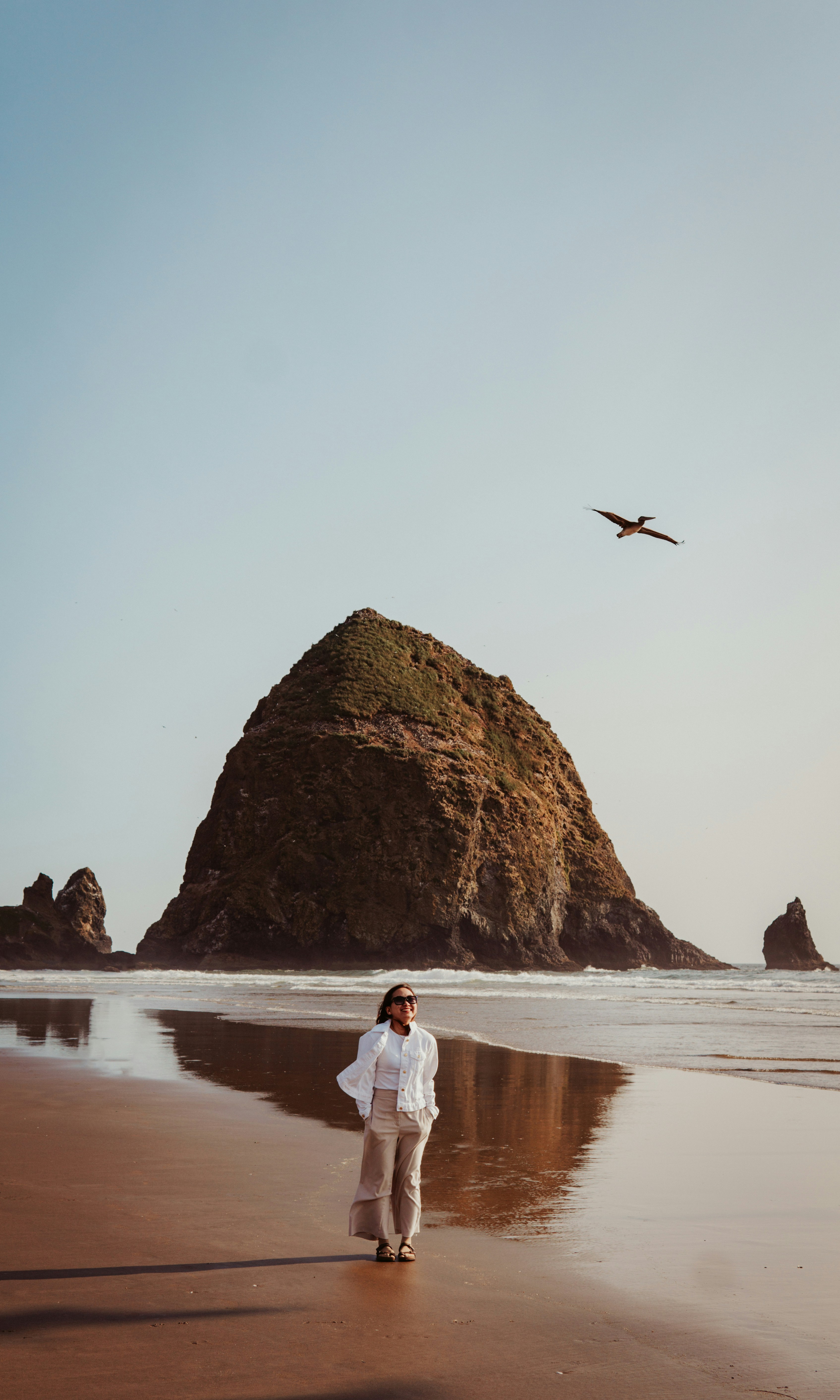 Woman walks on a beach with a large rock behind her.