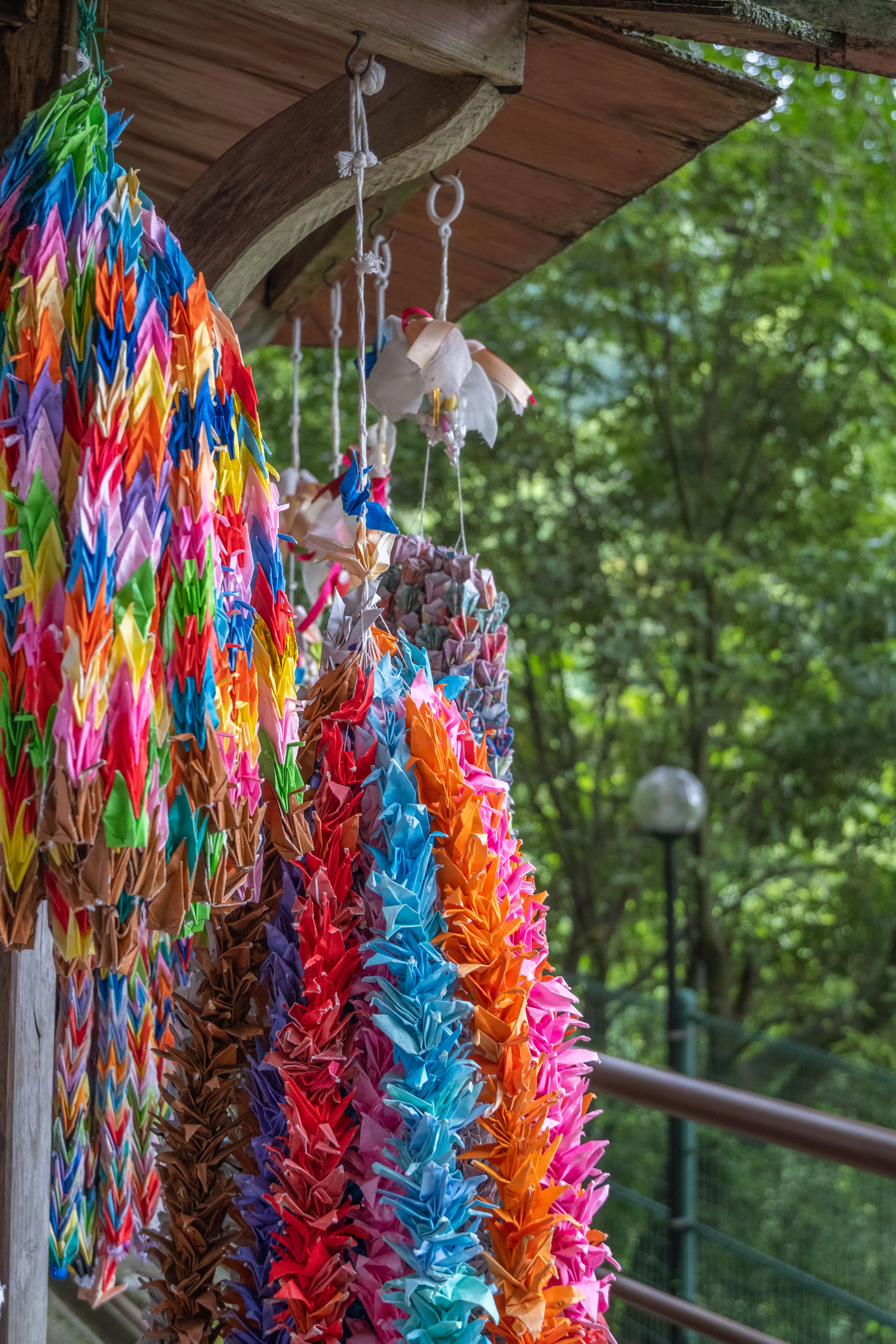 Rows of colorful folded paper cranes hang from a shrine eave against green trees.