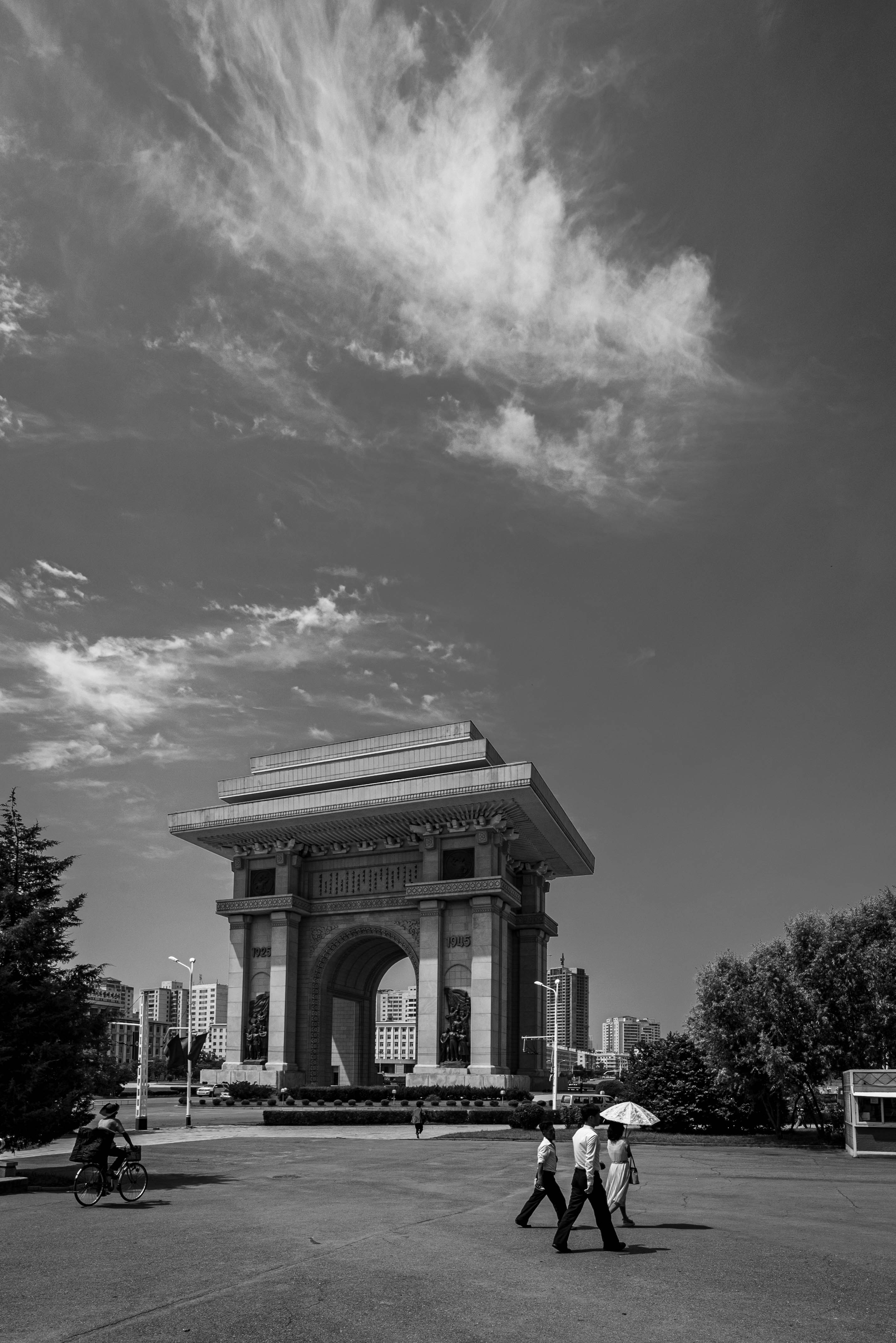People walk near a grand archway under a cloudy sky.