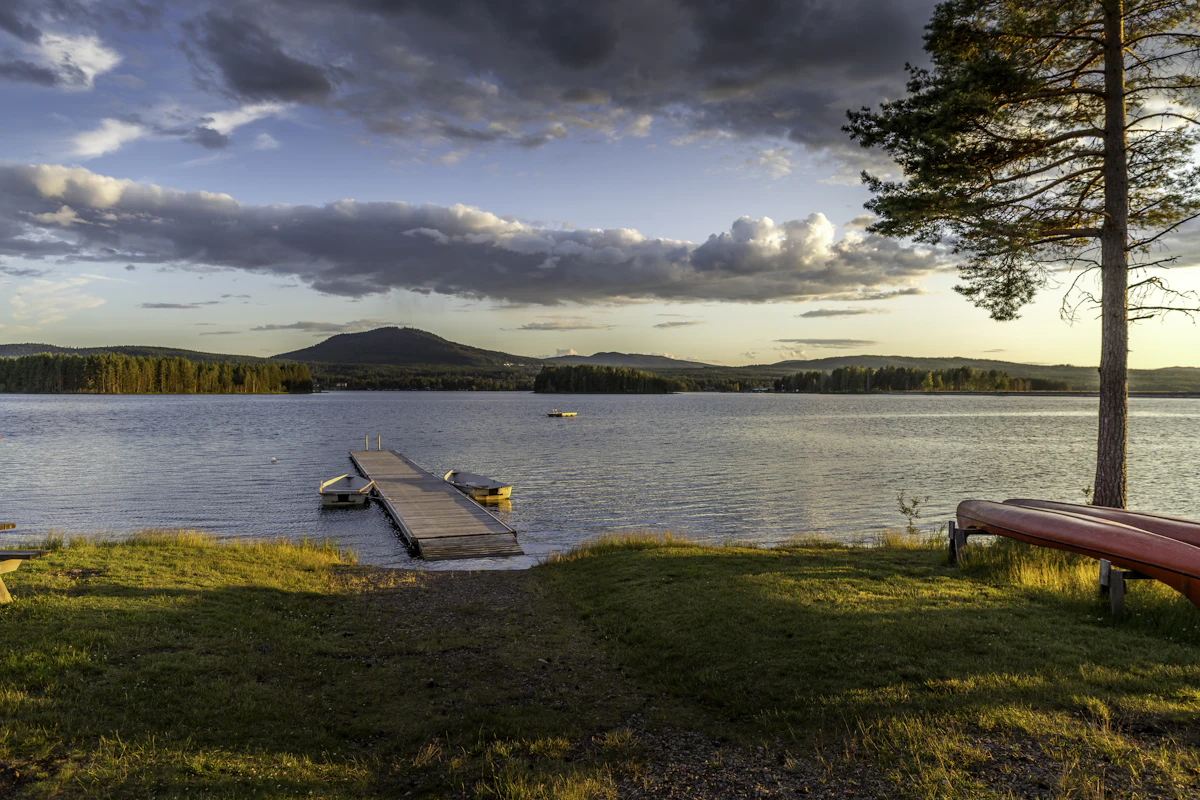 A jetty and canoes on a lake near Mora at sunset, with a pine on the shore and low hills behind