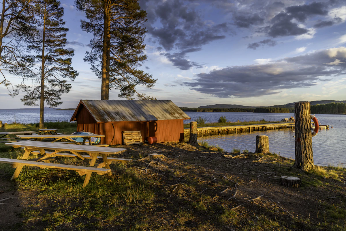 A red wooden shack and picnic tables under tall pines on a lakeshore near Mora