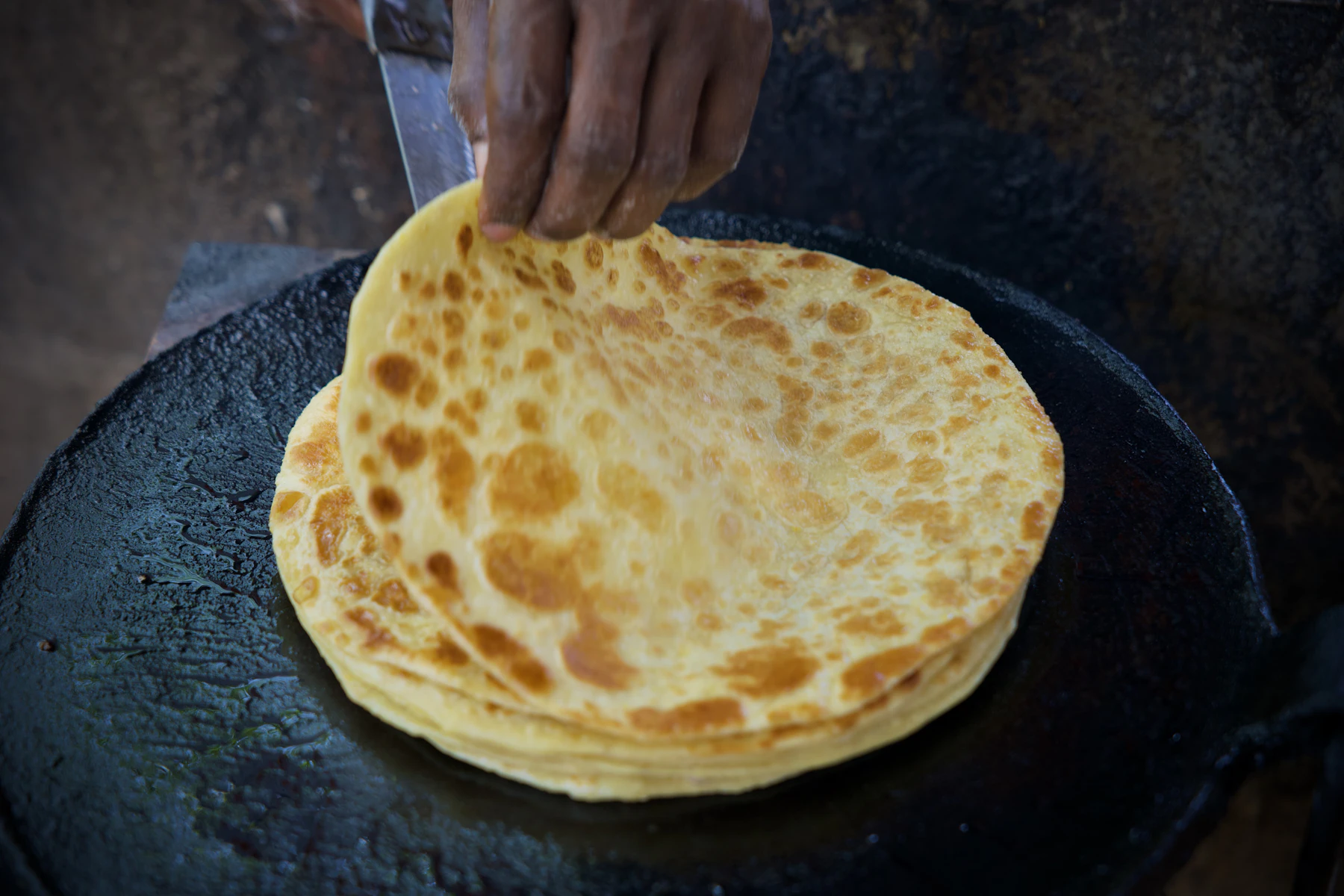 A hand lifting a browned flatbread from a stack cooking on a black griddle
