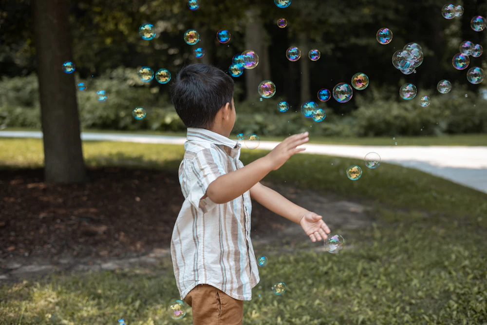 A young child in a garden reaching up towards a stream of floating soap bubbles