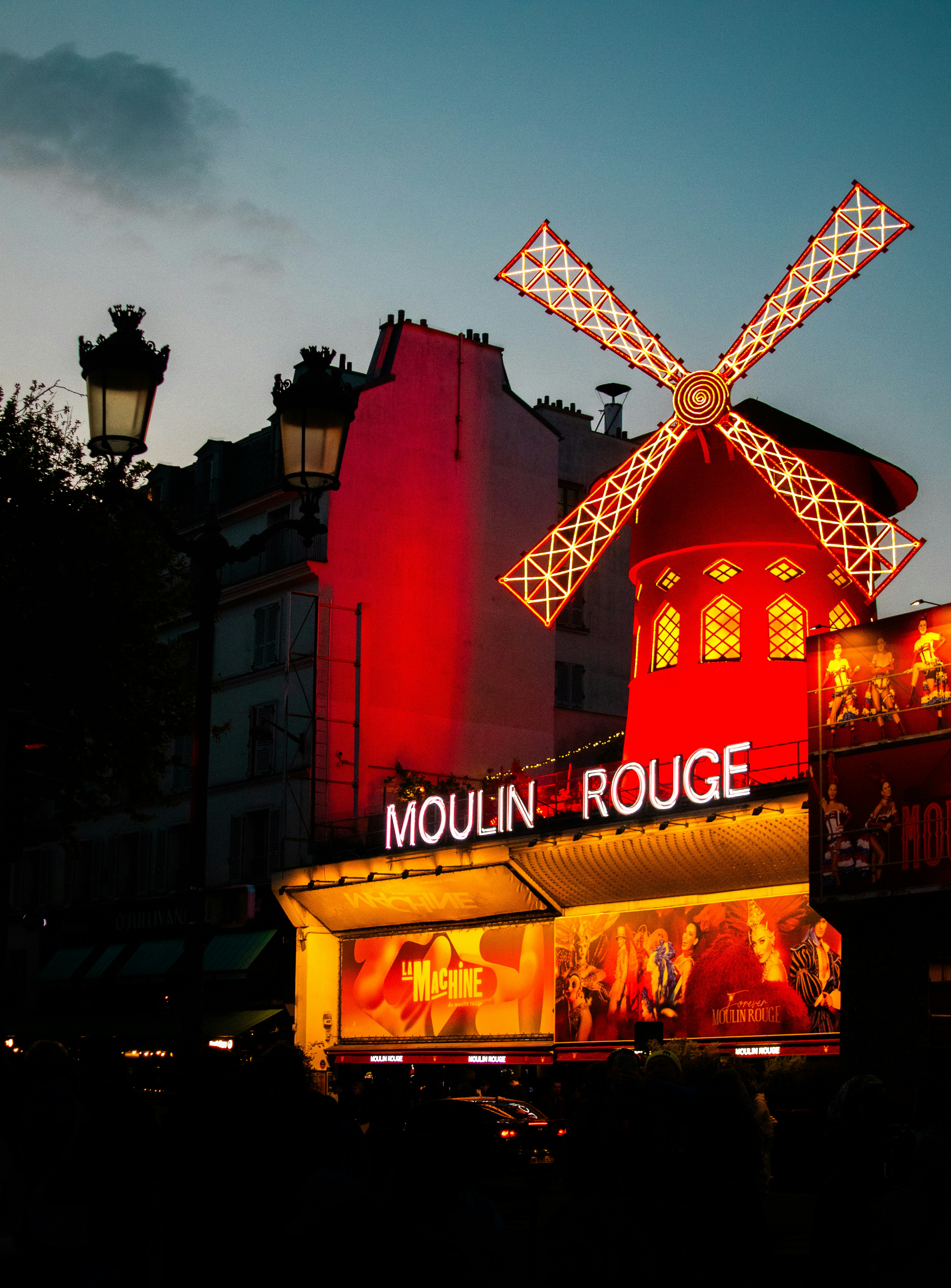 Vista ao crepúsculo do exterior do cabaré Moulin Rouge com o moinho vermelho iluminado acima da multidão em Paris, França.