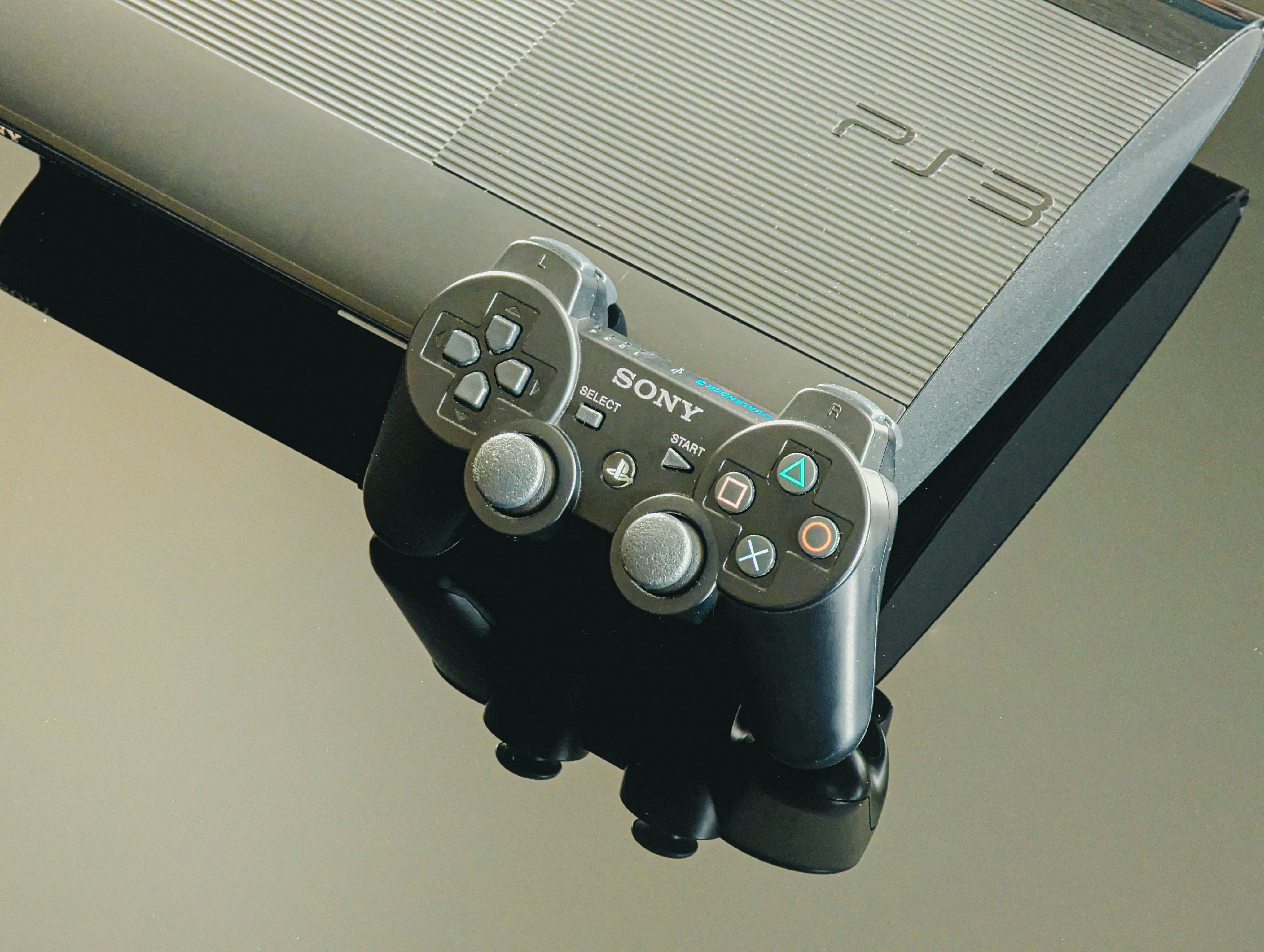 A highly reflective close-up of a PlayStation 3 (PS3) console and its wireless controller. The black plastic of the console and controller is set on a mirrored surface, highlighting the gaming theme, technology, and modern entertainment.