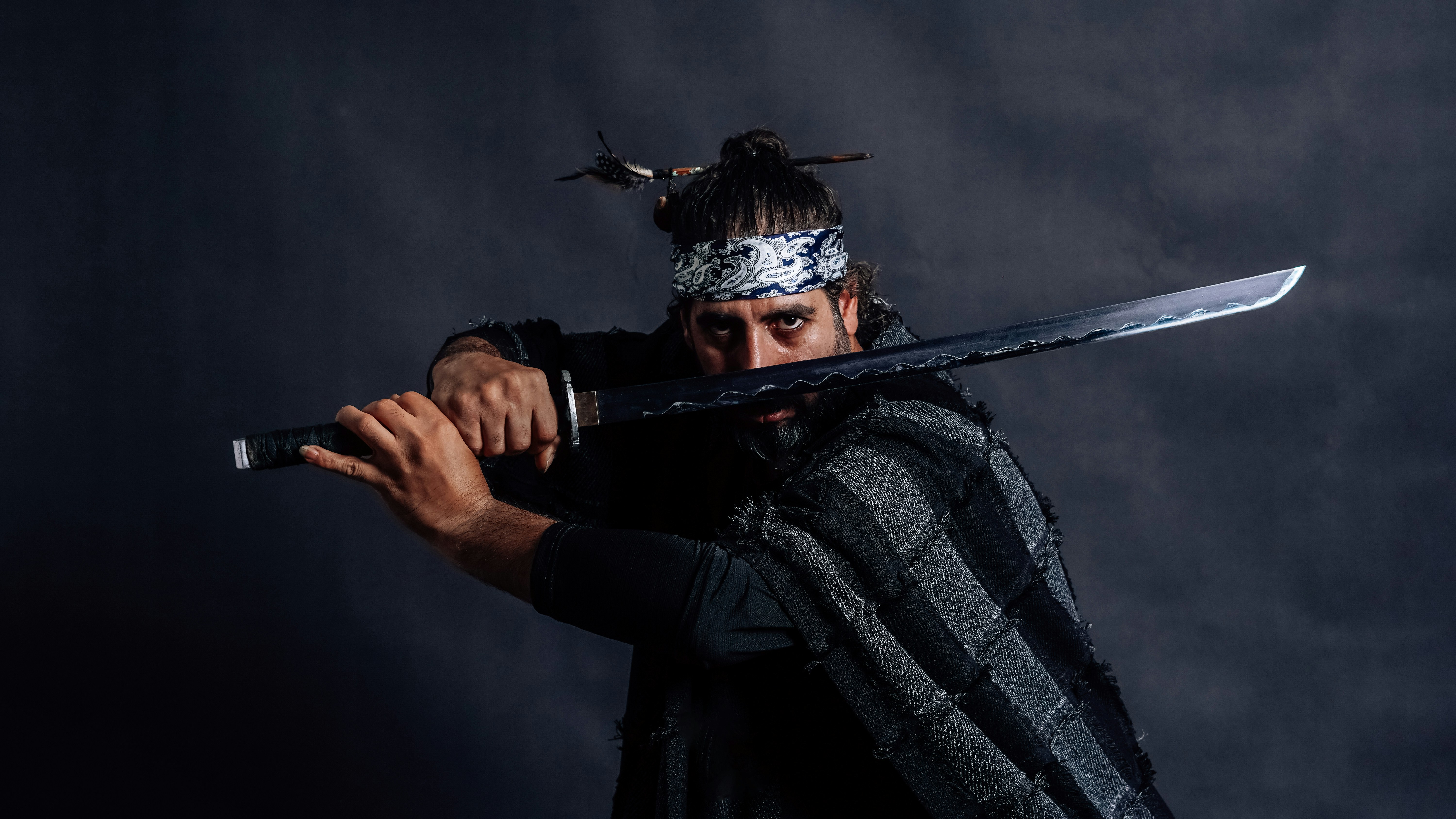 A high-resolution portrait of a bearded man dressed in traditional Japanese attire, holding a katana sword in a samurai stance, against a dark background. Dramatic lighting enhances the warrior aesthetic, blending historical Japanese culture with modern portrait photography.