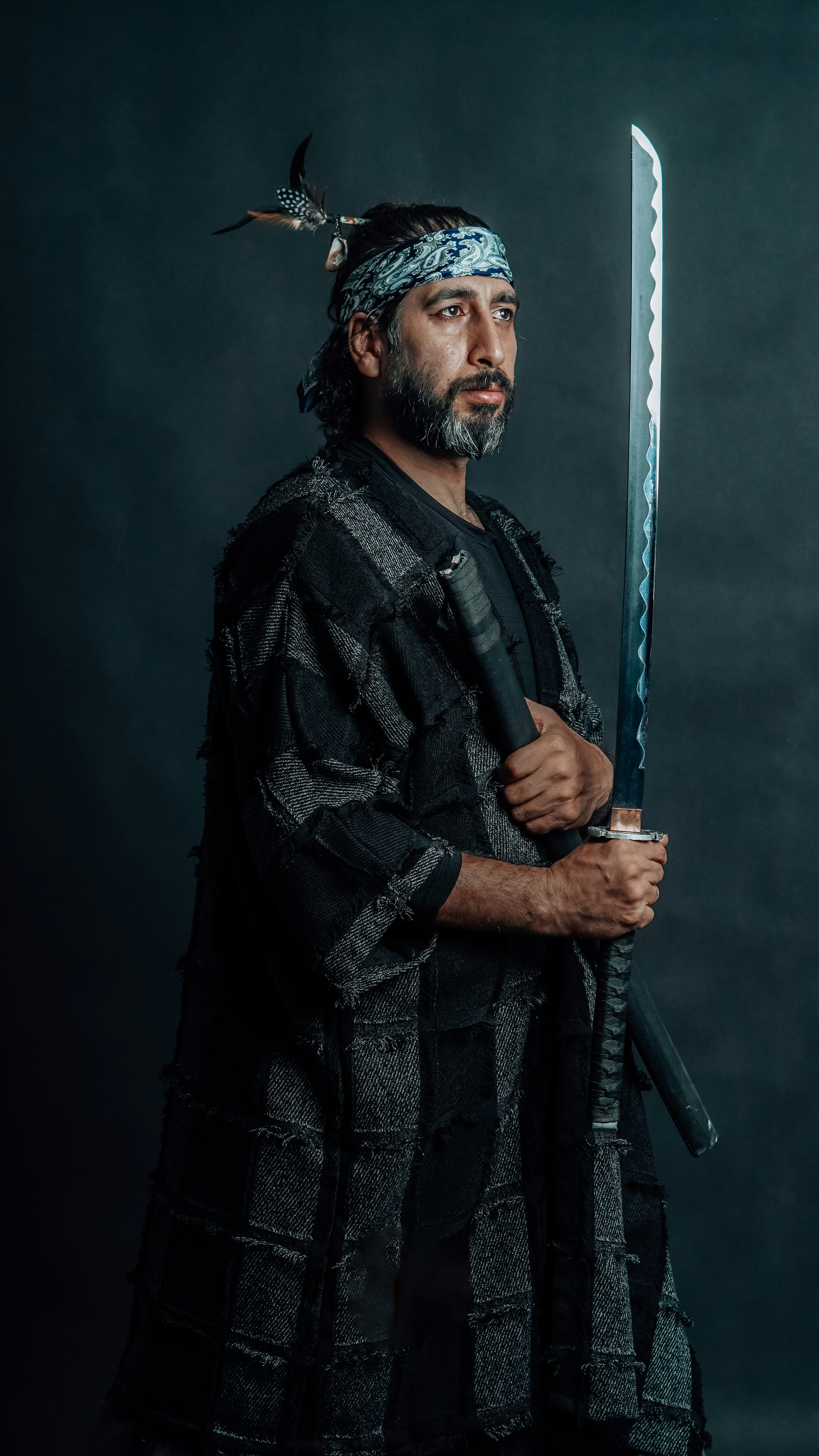 A high-resolution portrait of a bearded man dressed in traditional Japanese attire, holding a katana sword in a samurai stance, against a dark background. Dramatic lighting enhances the warrior aesthetic, blending historical Japanese culture with modern portrait photography.