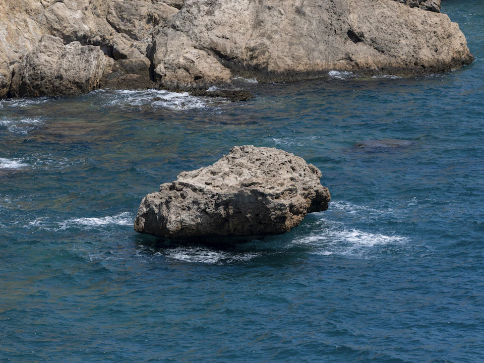 Large, light brown rock protrudes from rocky coastline into the deep blue ocean waters