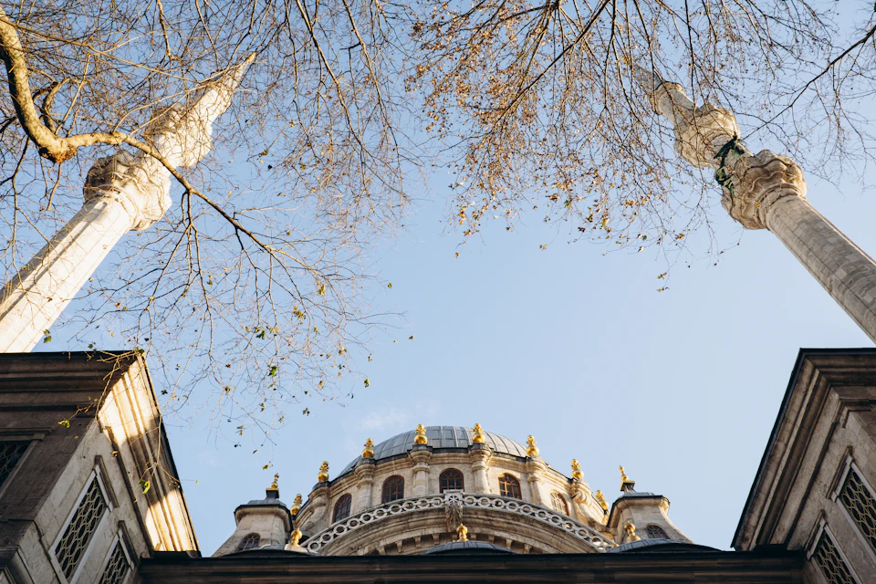 The image depicts a majestic white mosque with two towering minarets, one on each side, reaching towards the clear blue sky. The mosque's dome is adorned with intricate patterns and designs, adding to its grandeur. The perspective of the photo is from below, looking up at the mosque, emphasizing its height and architectural beauty. In the foreground, there are trees with bare branches, suggesting a colder season or location.