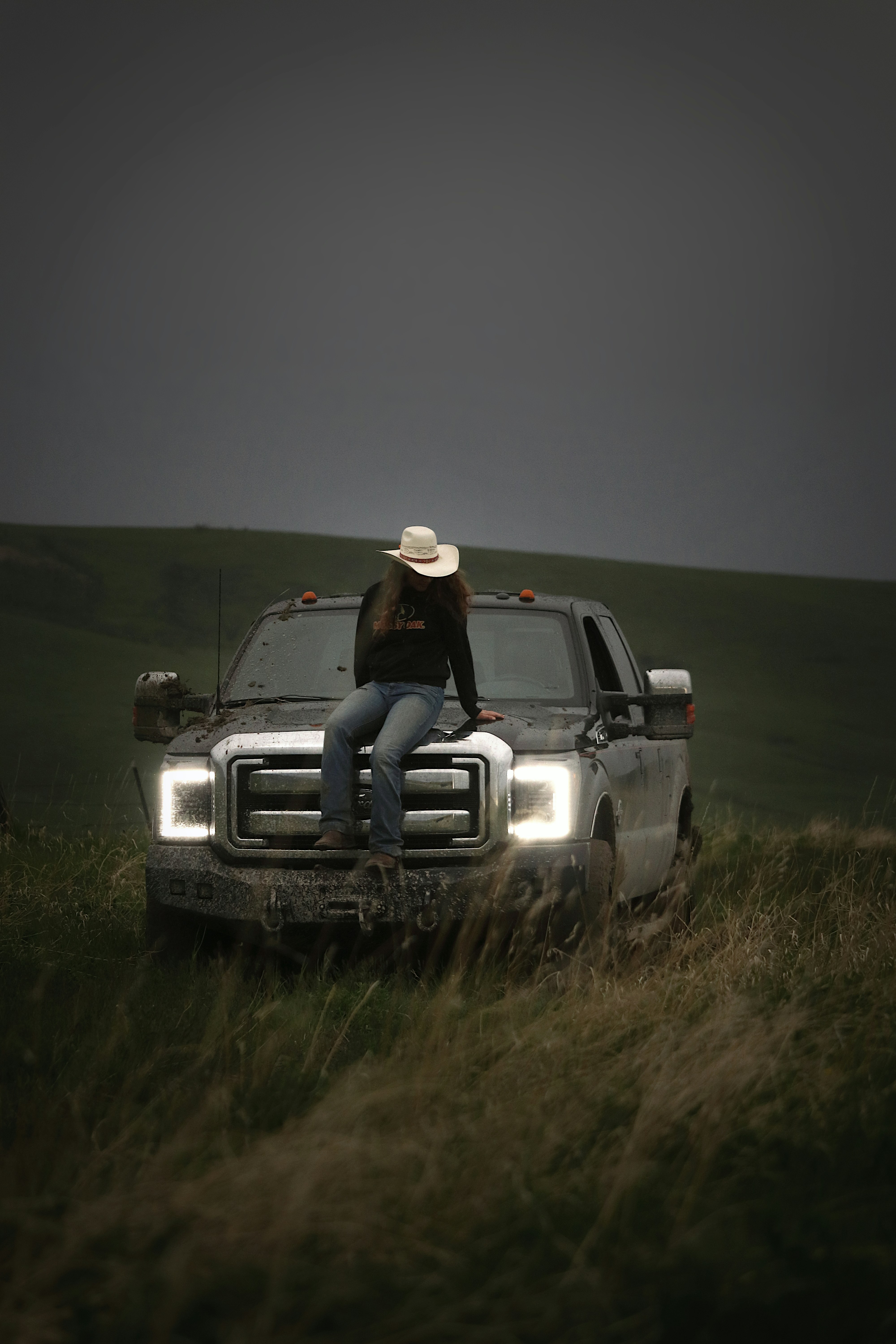 Cowgirl sits on a truck, under a cloudy sky.