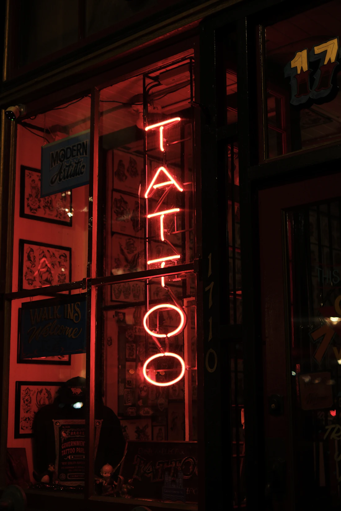 Red neon tattoo sign glowing in a dark studio window, flash sheets on the wall