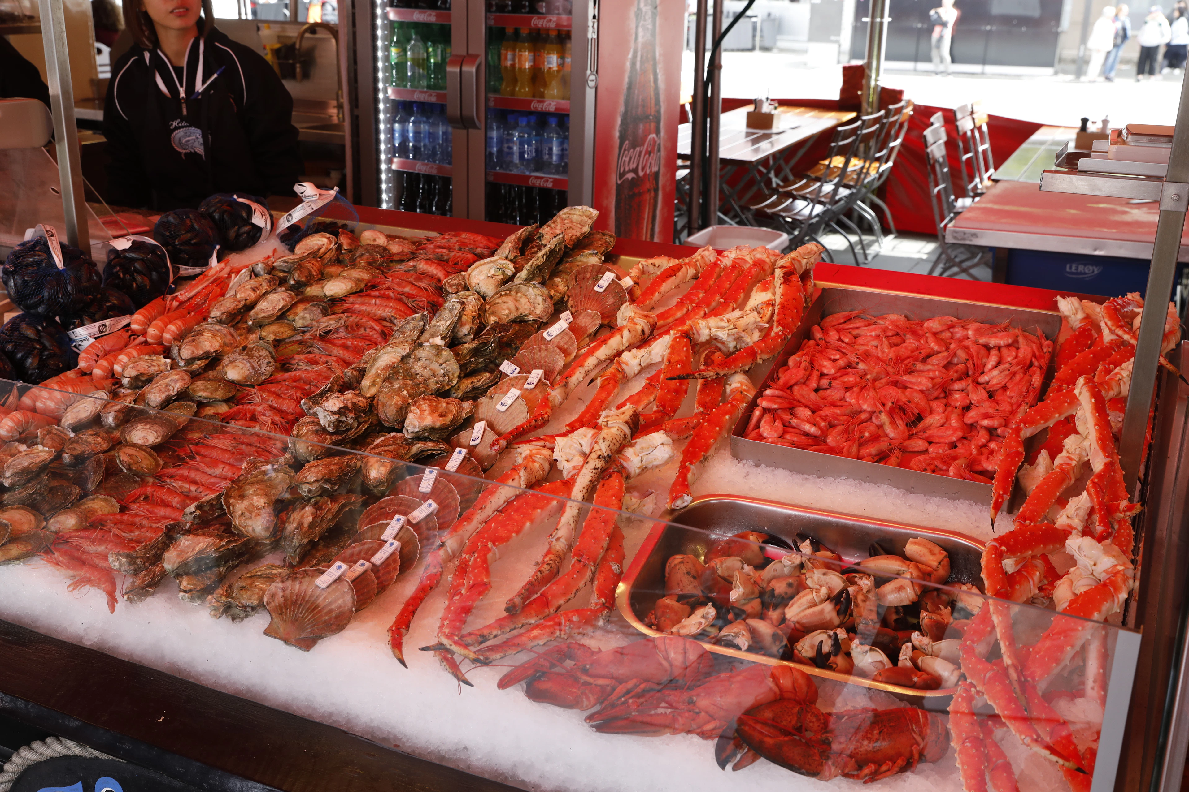 A Norwegian quayside fish-market stall banked with crushed ice, displaying king crab legs, langoustines, whole crabs and peeled shrimp as a vendor works behind the counter