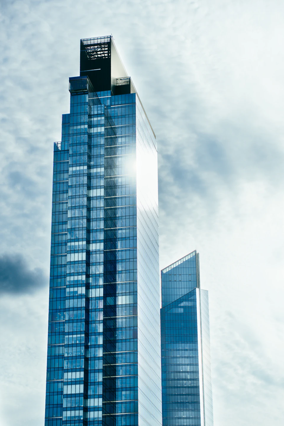 The image shows a tall, modern skyscraper with a glass facade that reflects the surrounding environment. The building is situated on a street corner and stands out against an overcast sky. The perspective of the photo is from below, looking up at the imposing height of the structure.