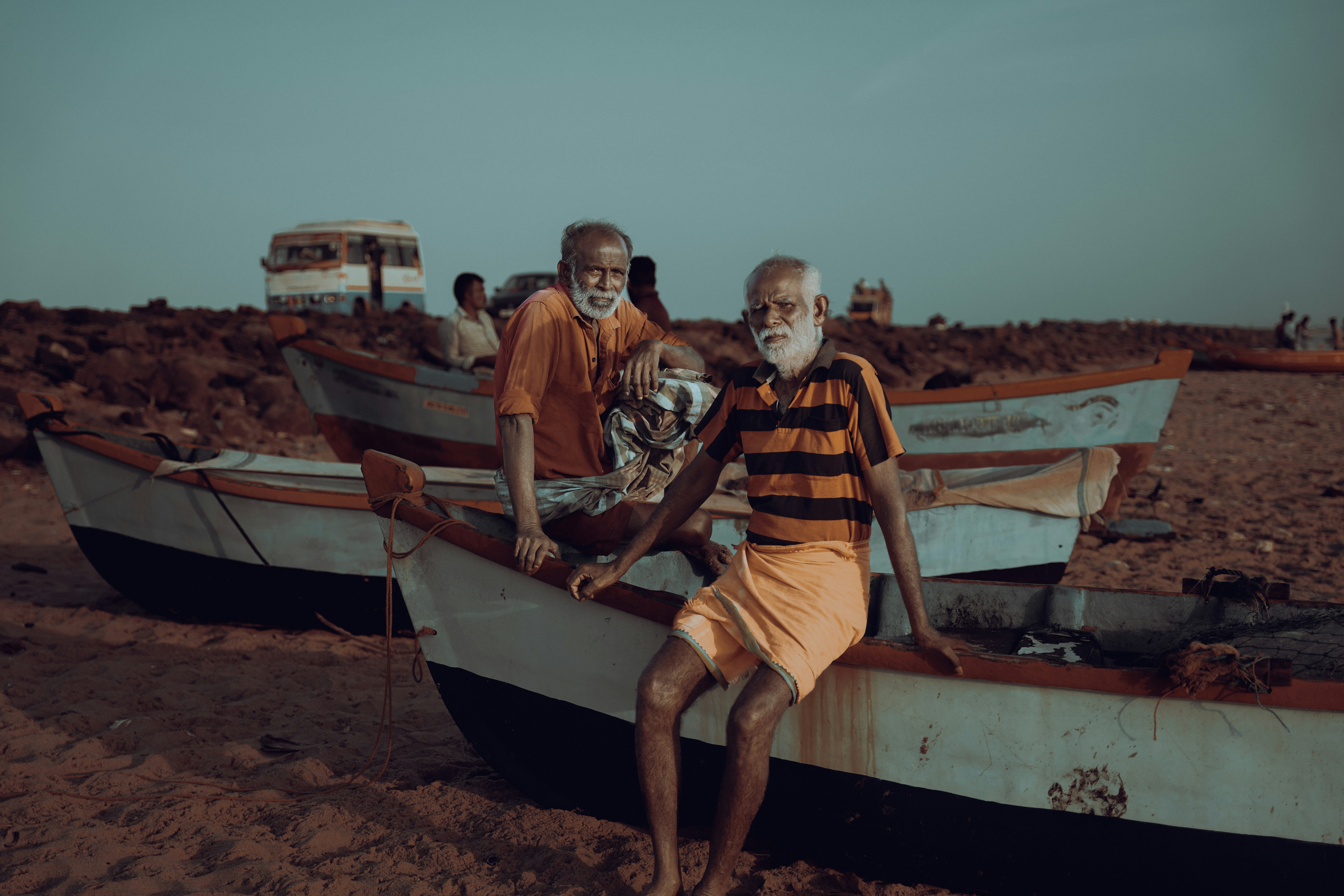 Two elderly men sit on boats on a beach.