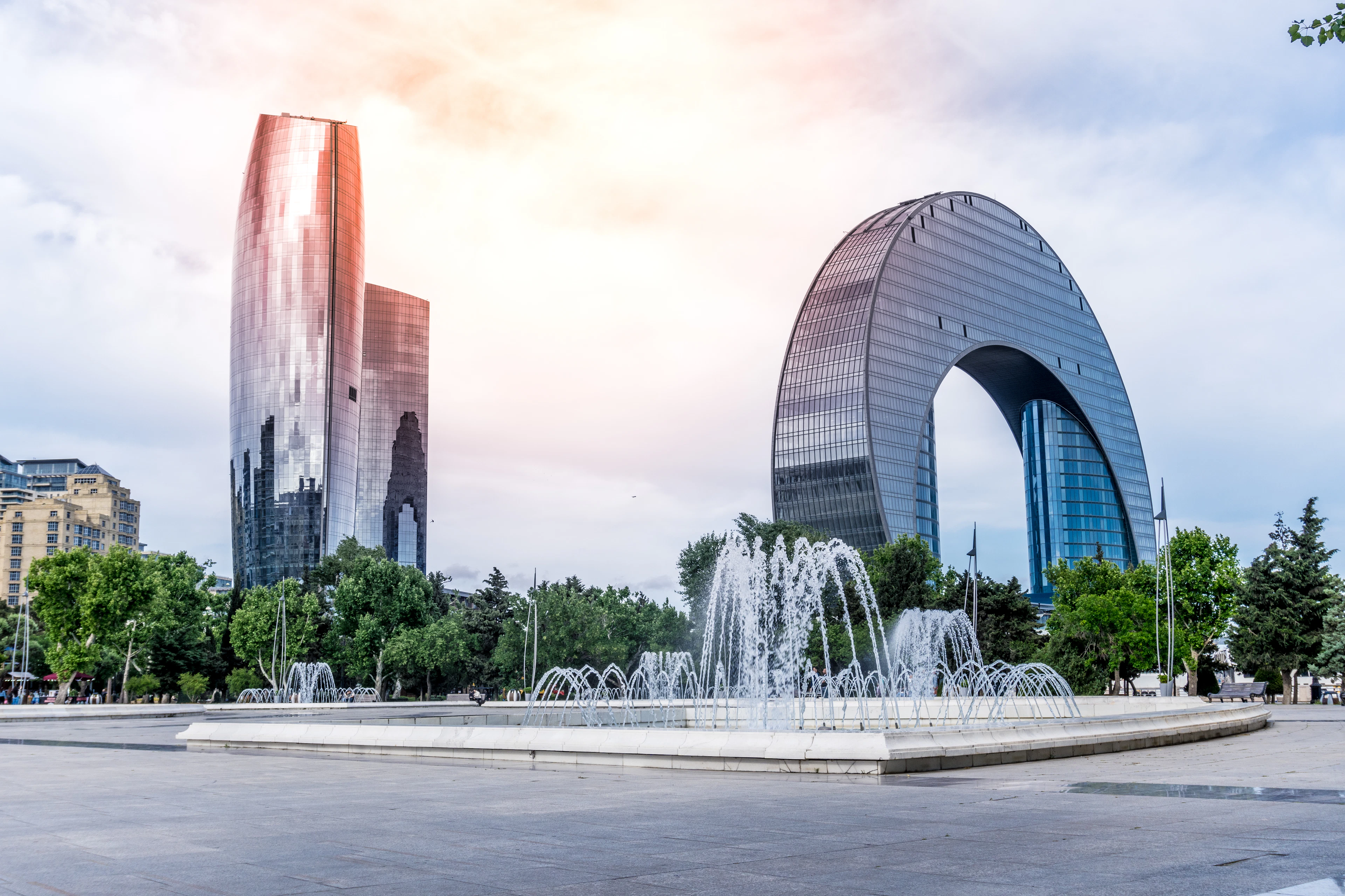 Fountains spray in a city square as sunlight glows behind a glass tower and an arched building.