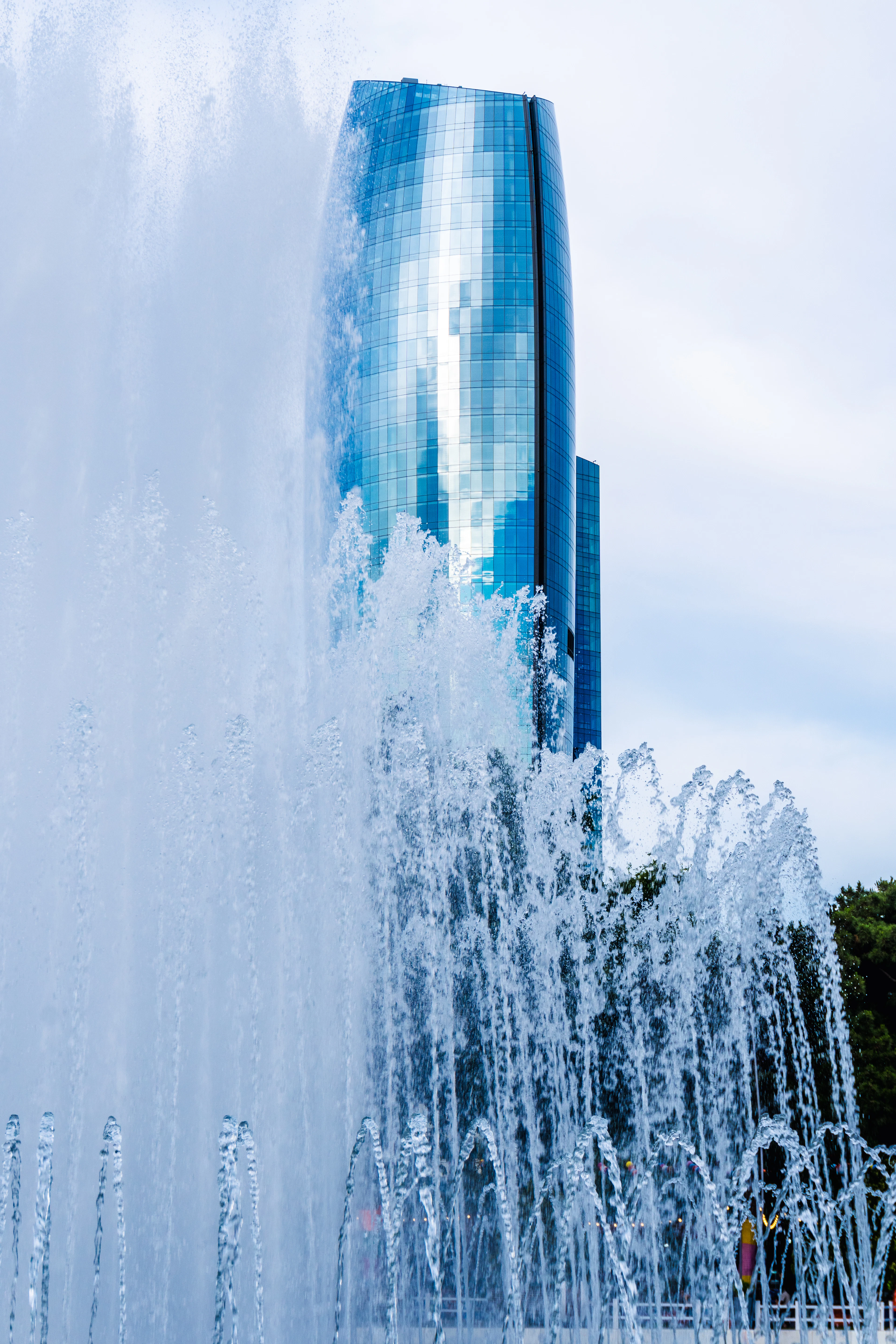 Fountain jets shoot up against a blue sky with a sail-shaped glass skyscraper behind them.