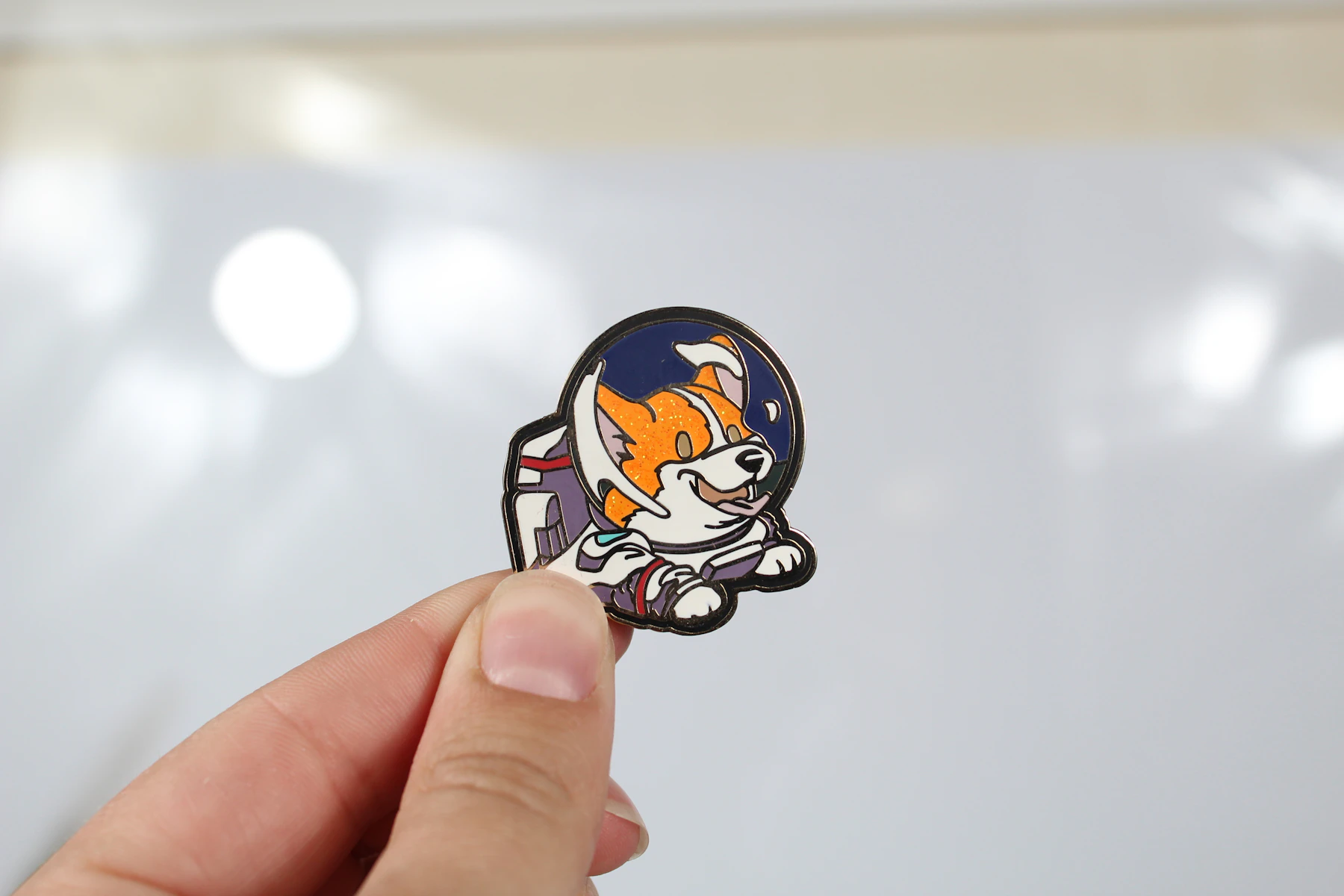Fingers holding up a single enamel pin of a corgi wearing a space helmet
