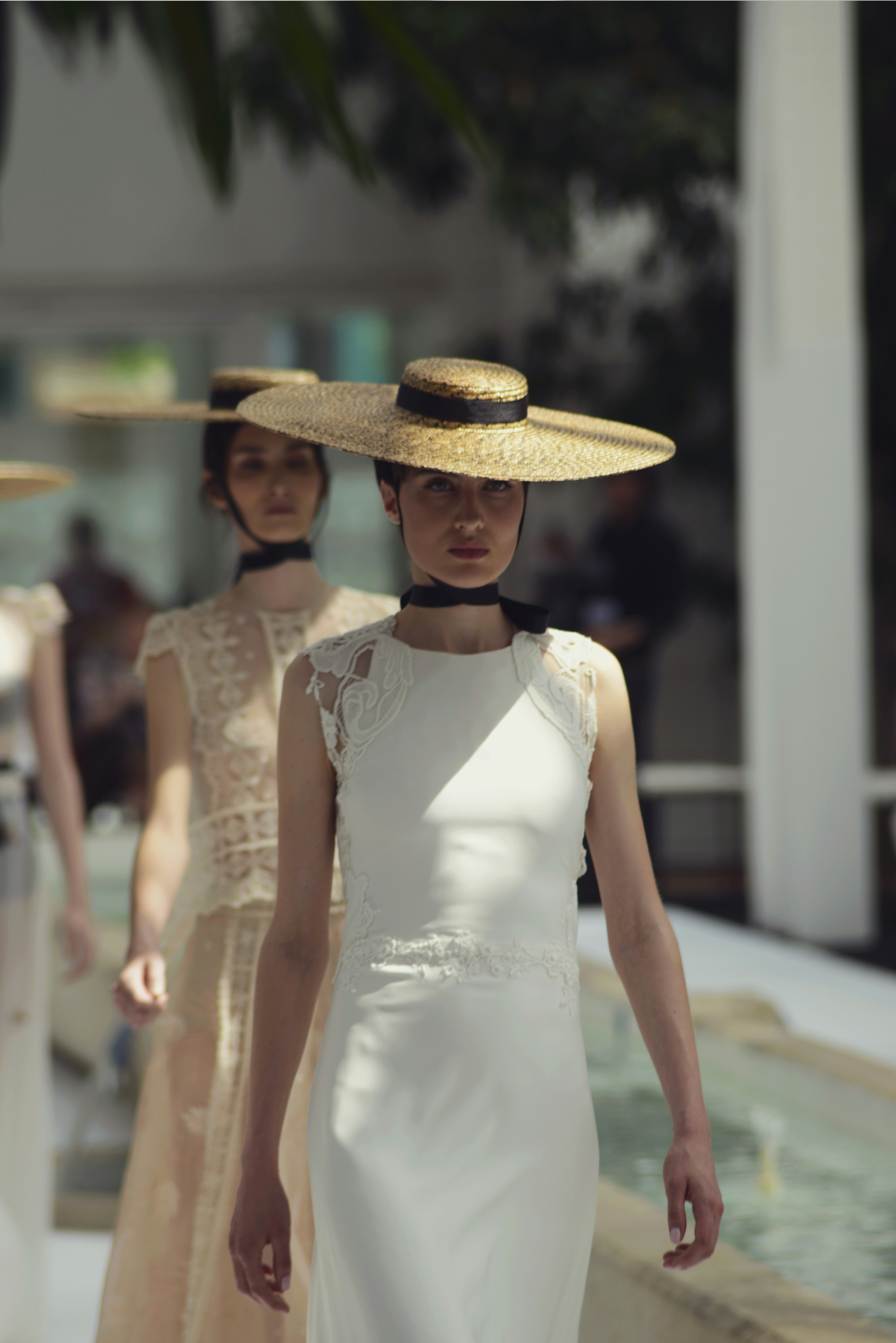 Two women wearing lace dresses and straw hats walk down a runway.