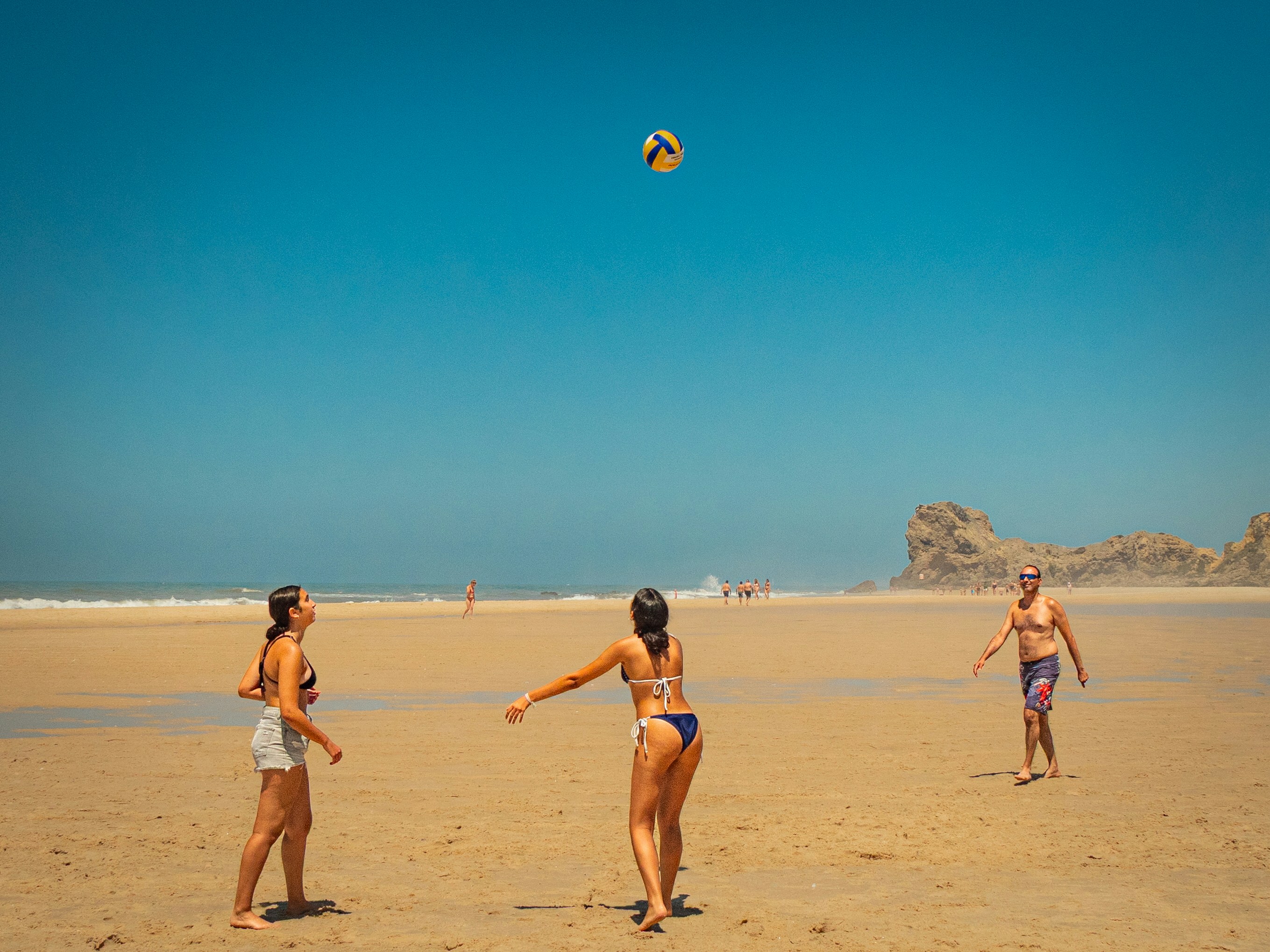 Minimal beach volleyball scene at Praia de Paredes da Vitória, Portugal—three players under a vast blue sky with the ball suspended above open sand and Atlantic cliffs.