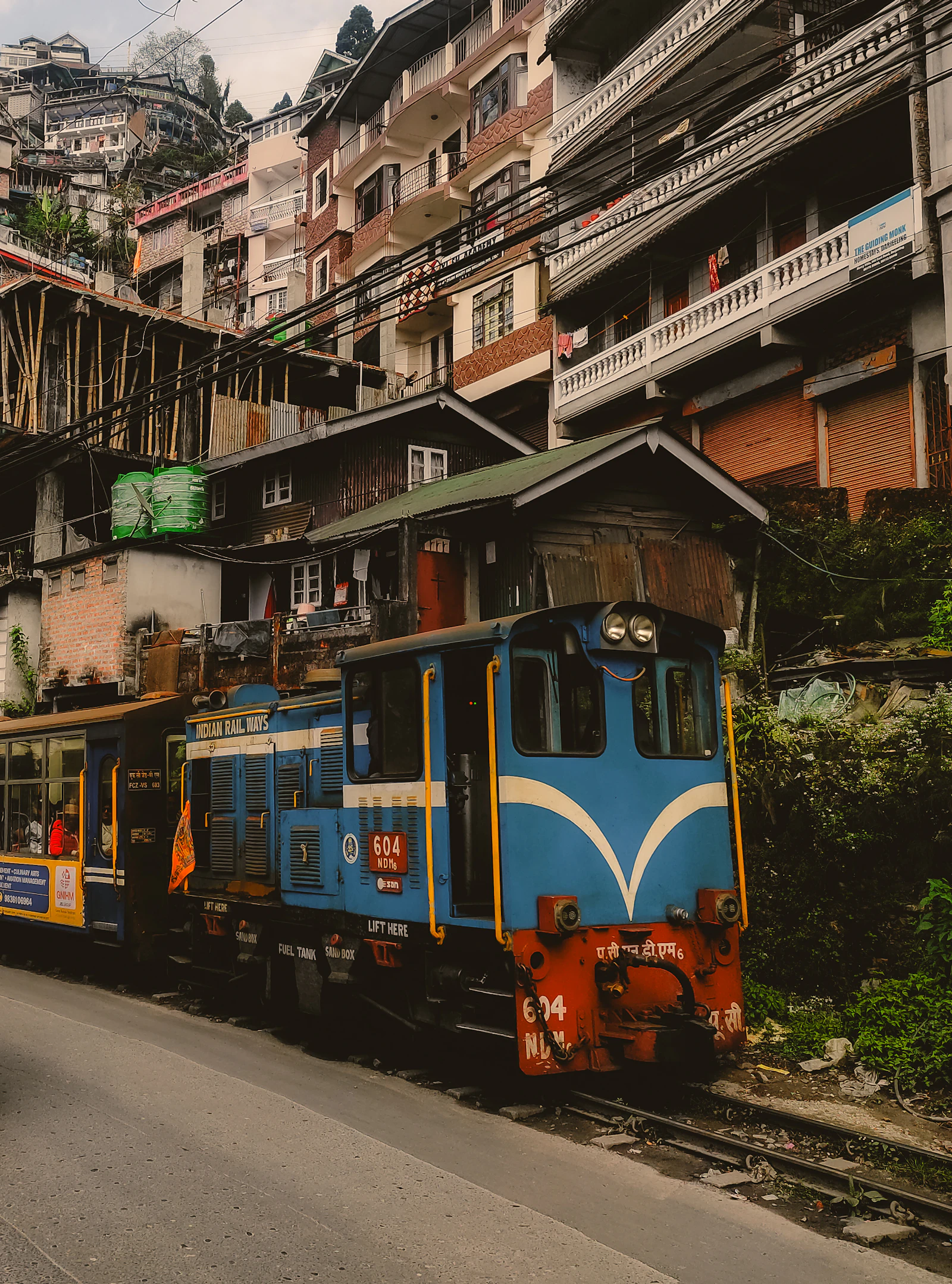 A blue toy train locomotive hauling carriages along the roadside track through a crowded hillside street in Darjeeling