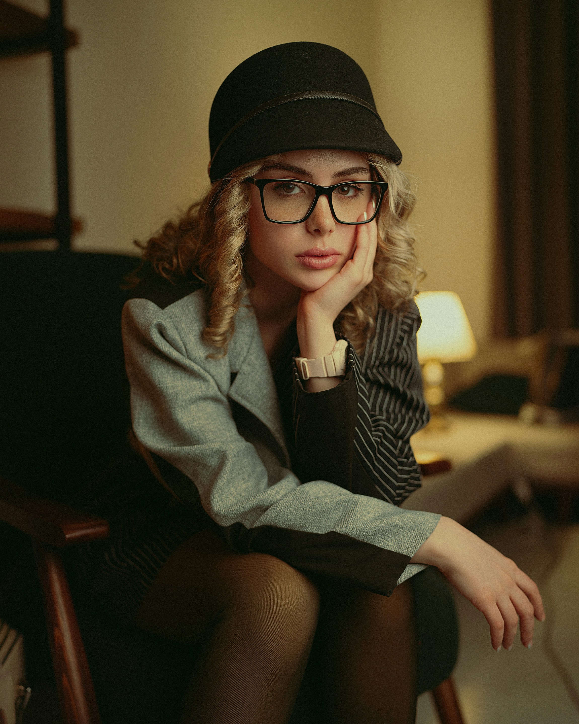 Une jeune femme avec des lunettes et un chapeau est assise pensivement.