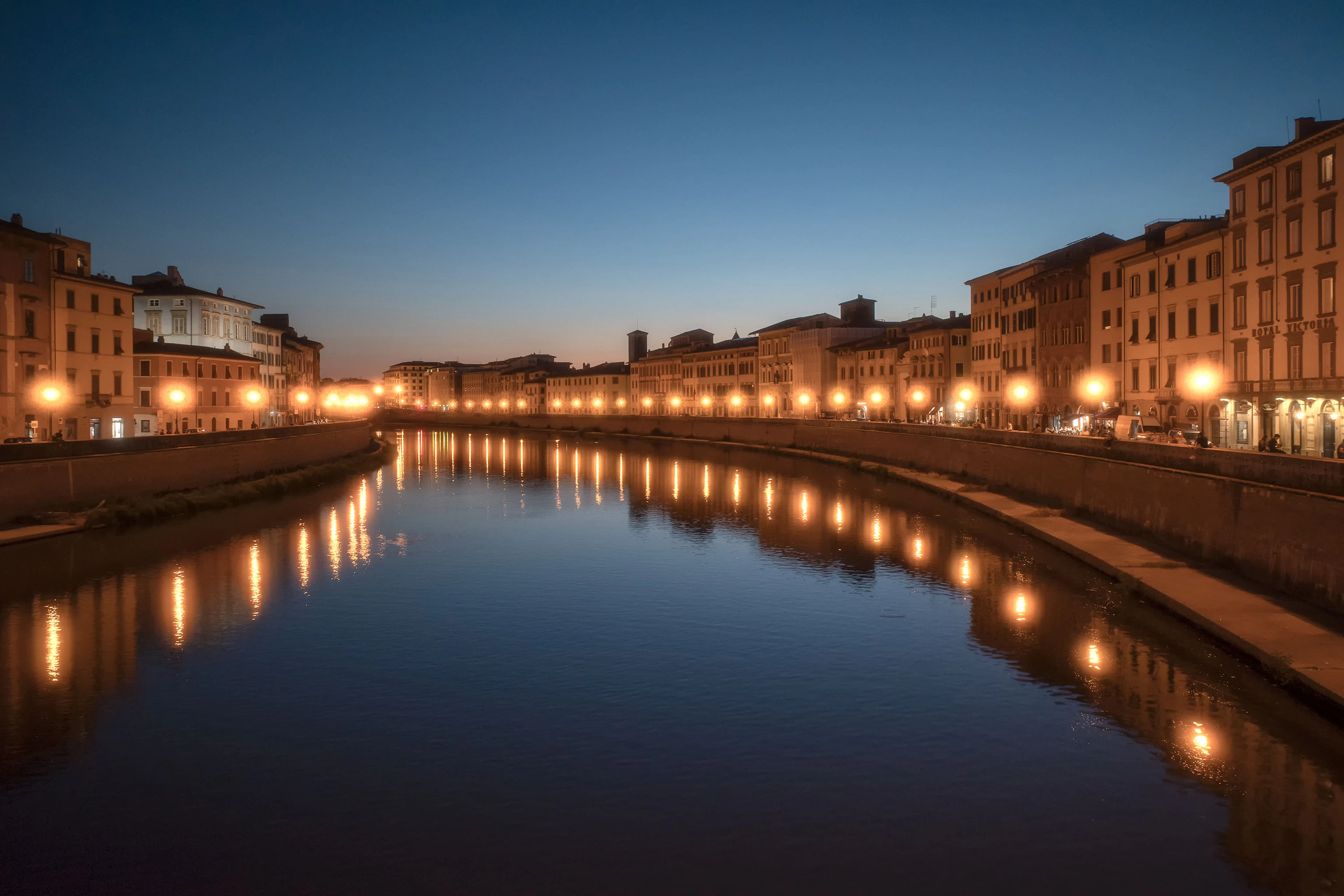 The Lungarni of Pisa at blue hour, two lines of street lamps running along the ochre palazzi of both embankments and doubling as long gold reflections in the still surface of the Arno.