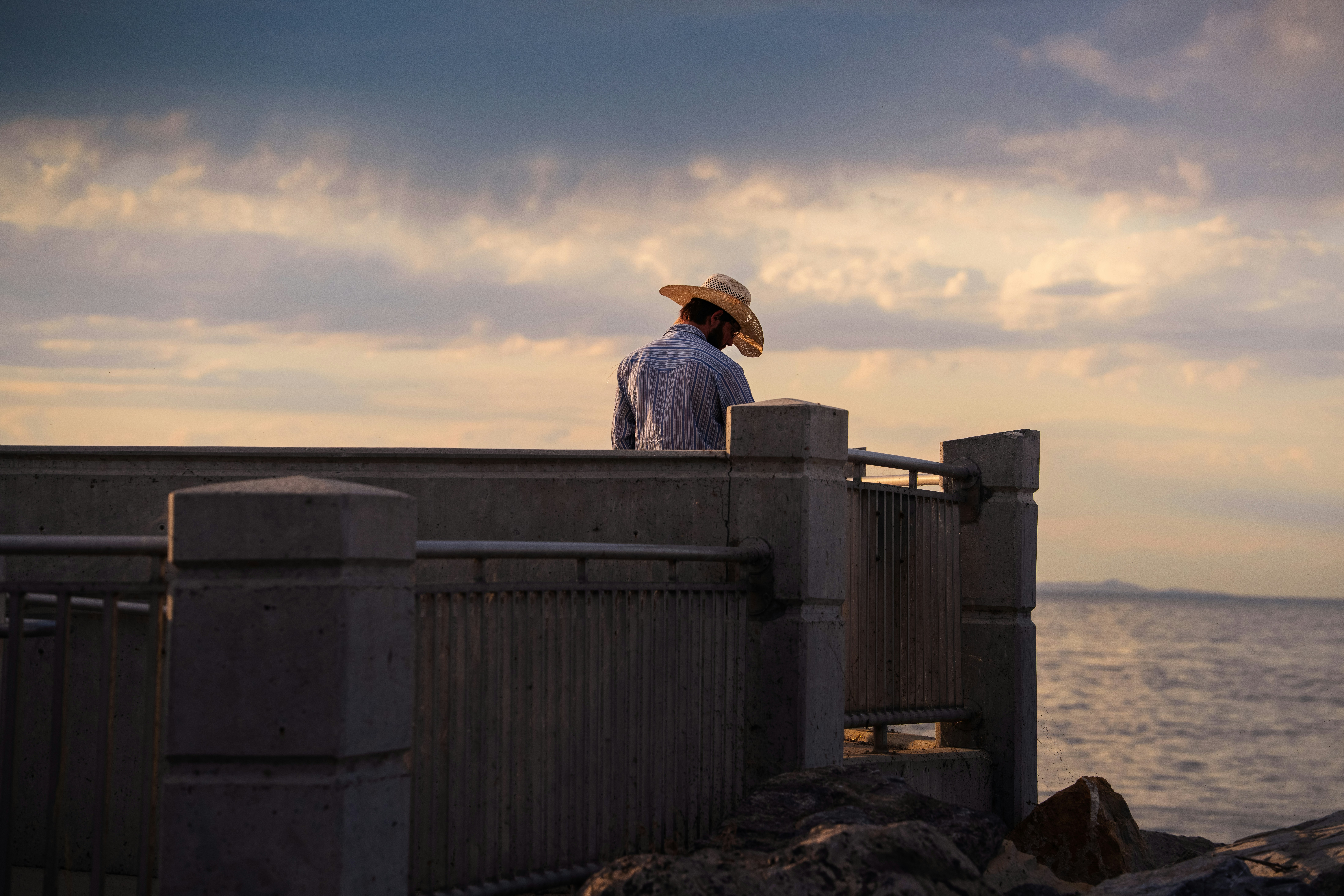 A man in a cowboy hat stands quietly on a concrete pier overlooking the Great Salt Lake during sunset. The warm light of the evening sky casts soft tones over the water and the structure, creating a peaceful, contemplative moment by the lakeshore.