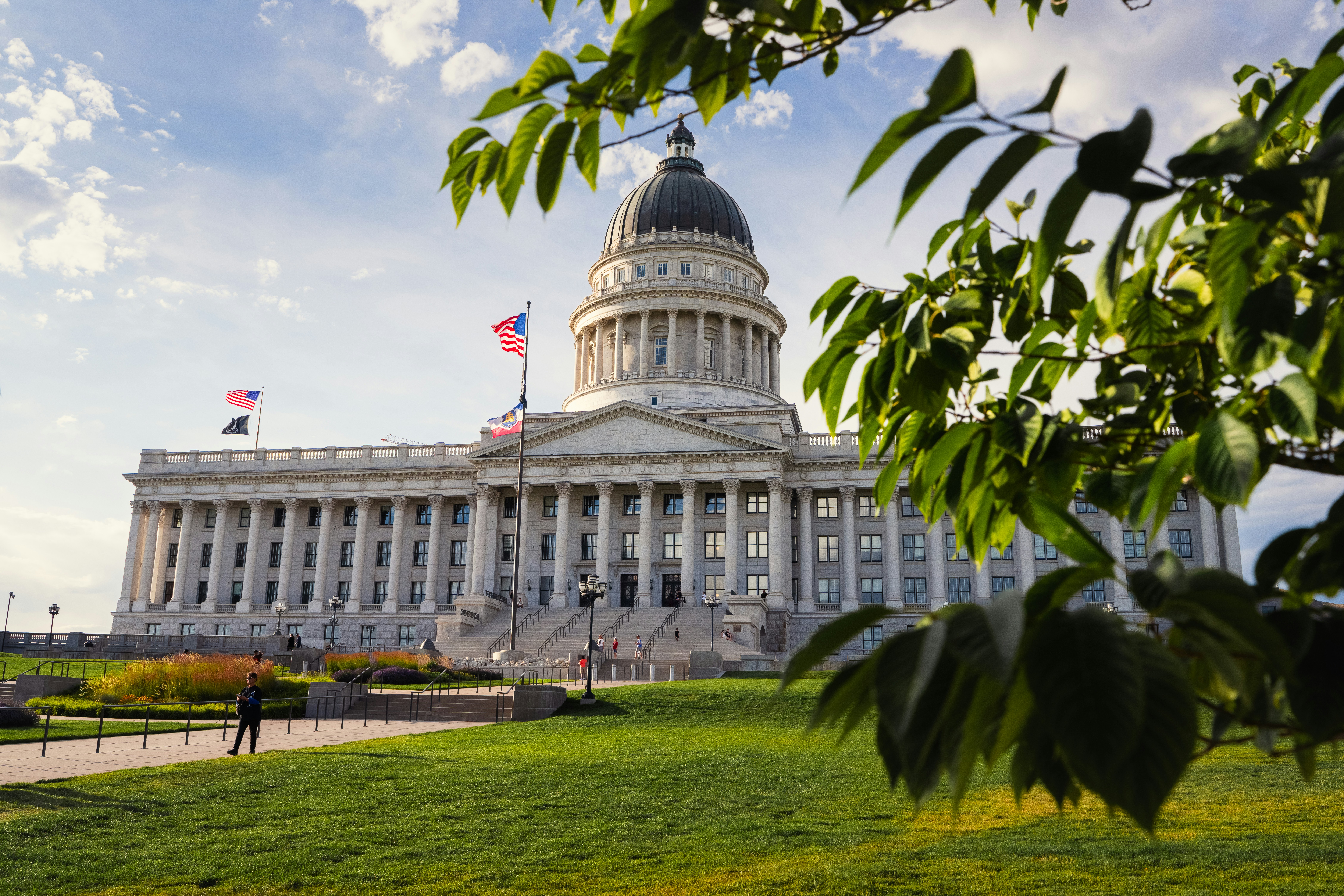 The Utah State Capitol building in Salt Lake City, framed by green tree leaves under a bright sky.