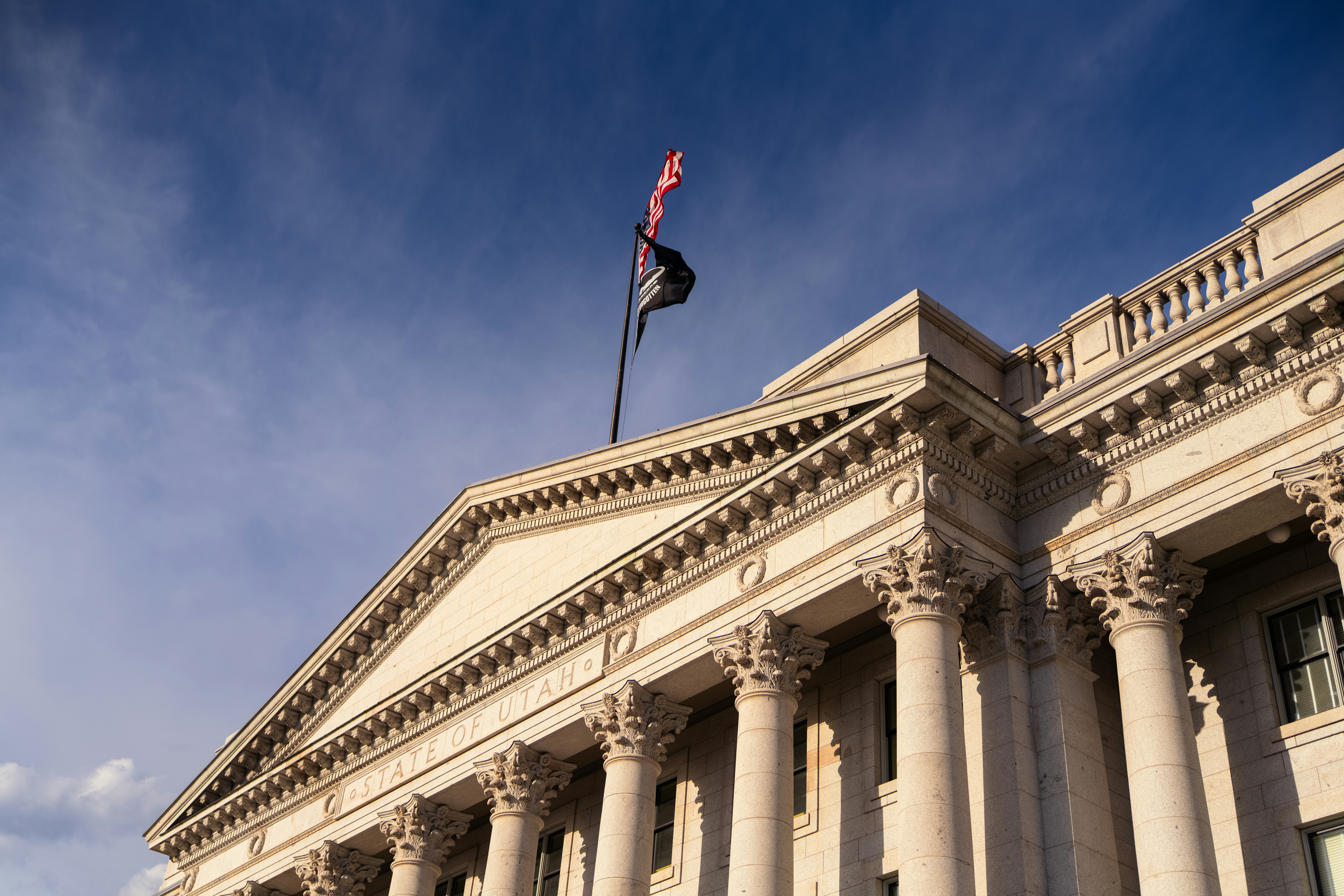 Close-up view of the Utah State Capitol's neoclassical facade in Salt Lake City, showcasing grand Corinthian columns, ornate stonework, and the American and POW/MIA flags flying above against a deep blue sky in warm evening light.