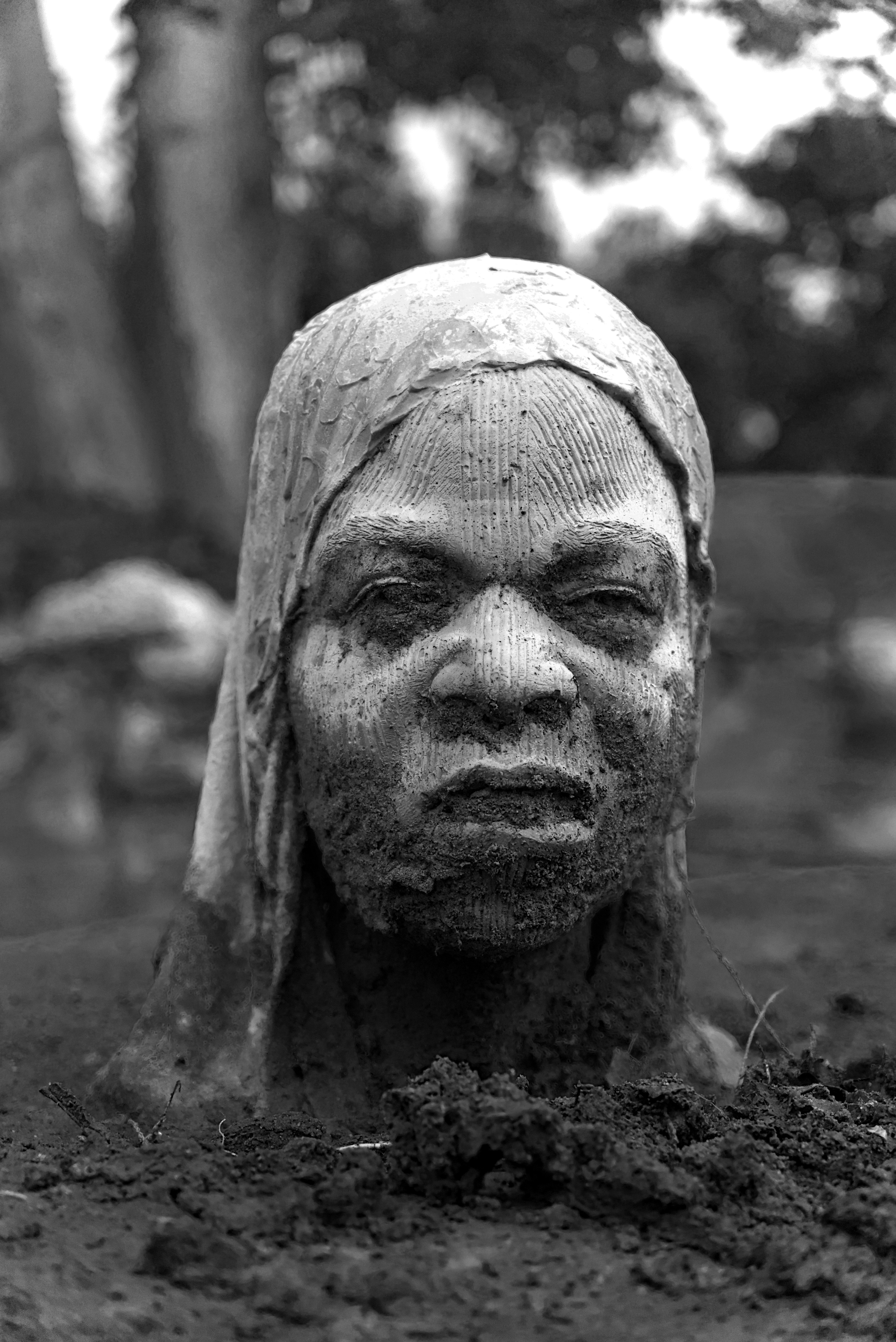 A head sculpture covered in mud emerges from the earth.