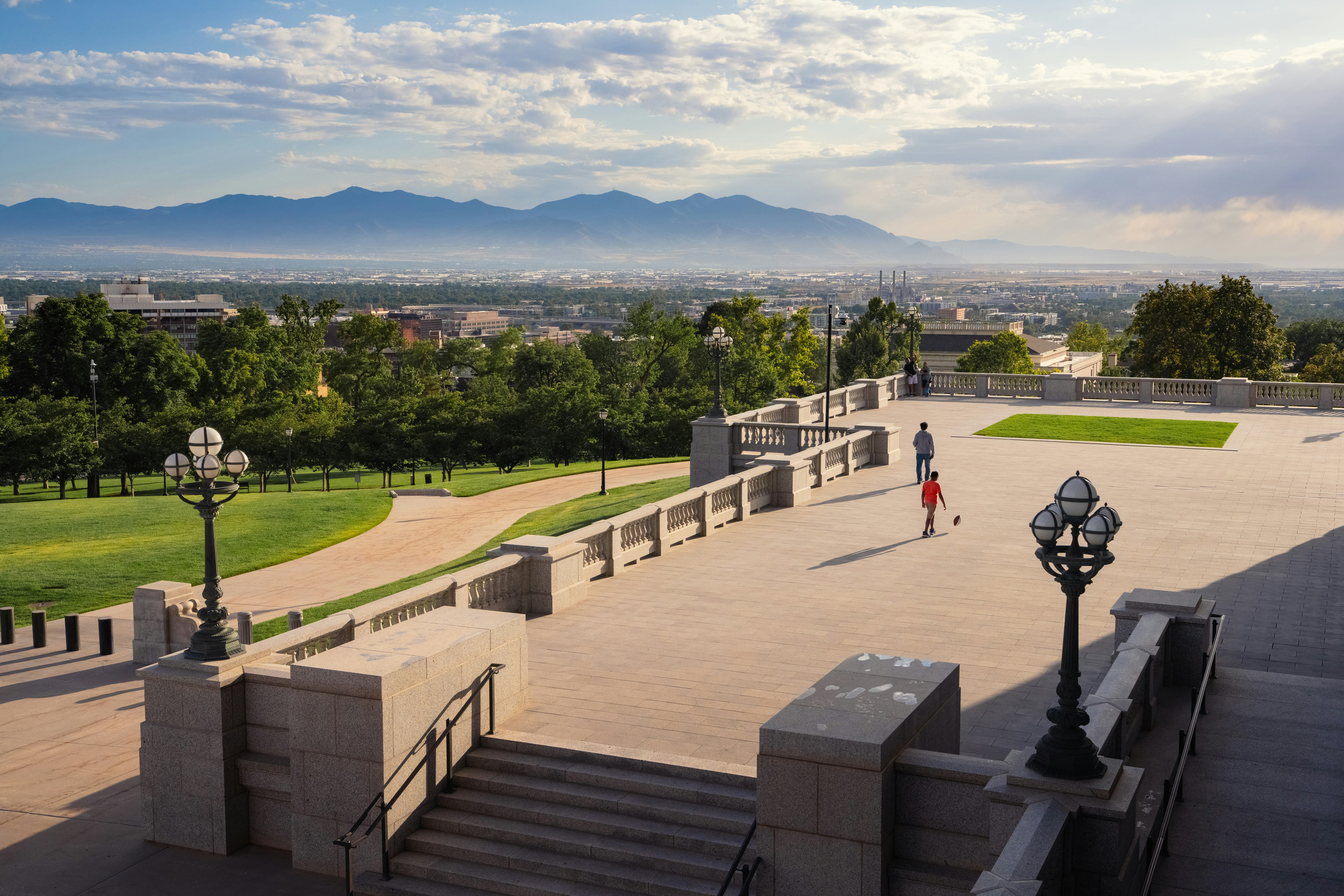 The terrace of the Utah State Capitol in Salt Lake City offers a sweeping view of the city skyline, framed by lush green lawns and tree-lined walkways. In the distance, the Wasatch Mountains rise under a hazy summer sky, with sunlight breaking through scattered clouds. A few people enjoy the open space, adding a sense of scale to the grand architecture and expansive landscape.