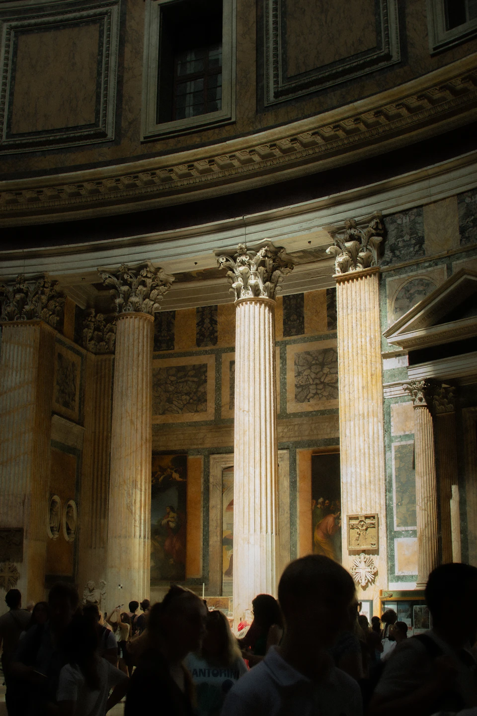 The image depicts a group of people gathered inside the Colosseum, an ancient Roman amphitheater located in Rome, Italy. The perspective is from within the structure, looking out onto the crowd. The Colosseum's architectural features are clearly visible, including three tall columns and a large archway at the center. These elements contribute to the grandeur of the scene. The people inside the Colosseum appear to be engaged in various activities, creating a lively atmosphere within the historic site.

The image is bathed in natural light streaming through windows on either side, highlighting the intricate details and textures of the stone walls and columns.