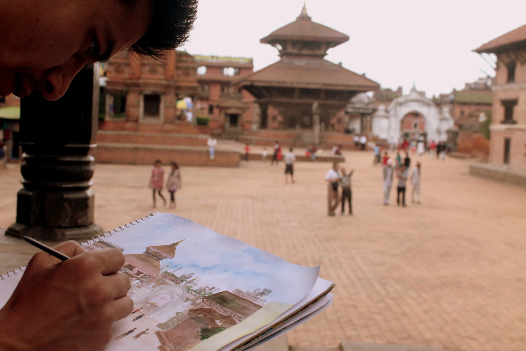 A hand sketching a temple in a sketchbook at the edge of a brick town square, with people walking across the square beyond