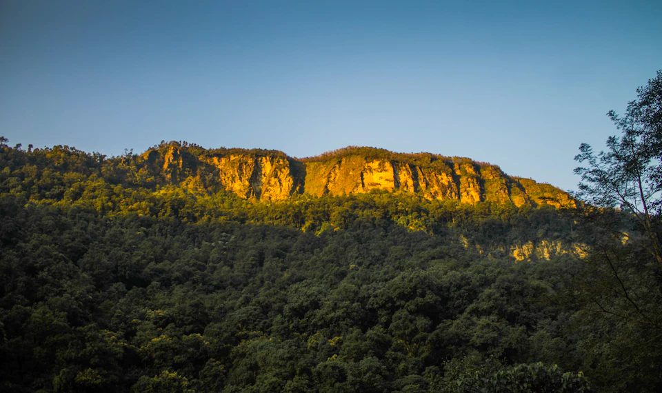 The image depicts a majestic mountain range with a large, light brown cliff face rising from the base to a darker brown peak. The mountain is bathed in warm sunlight, casting long shadows and highlighting its rugged texture. The sky above mirrors the color of the mountain's peak, creating a harmonious blend of colors. In the foreground, trees can be seen peeking through the foliage, adding depth and scale to the scene.
