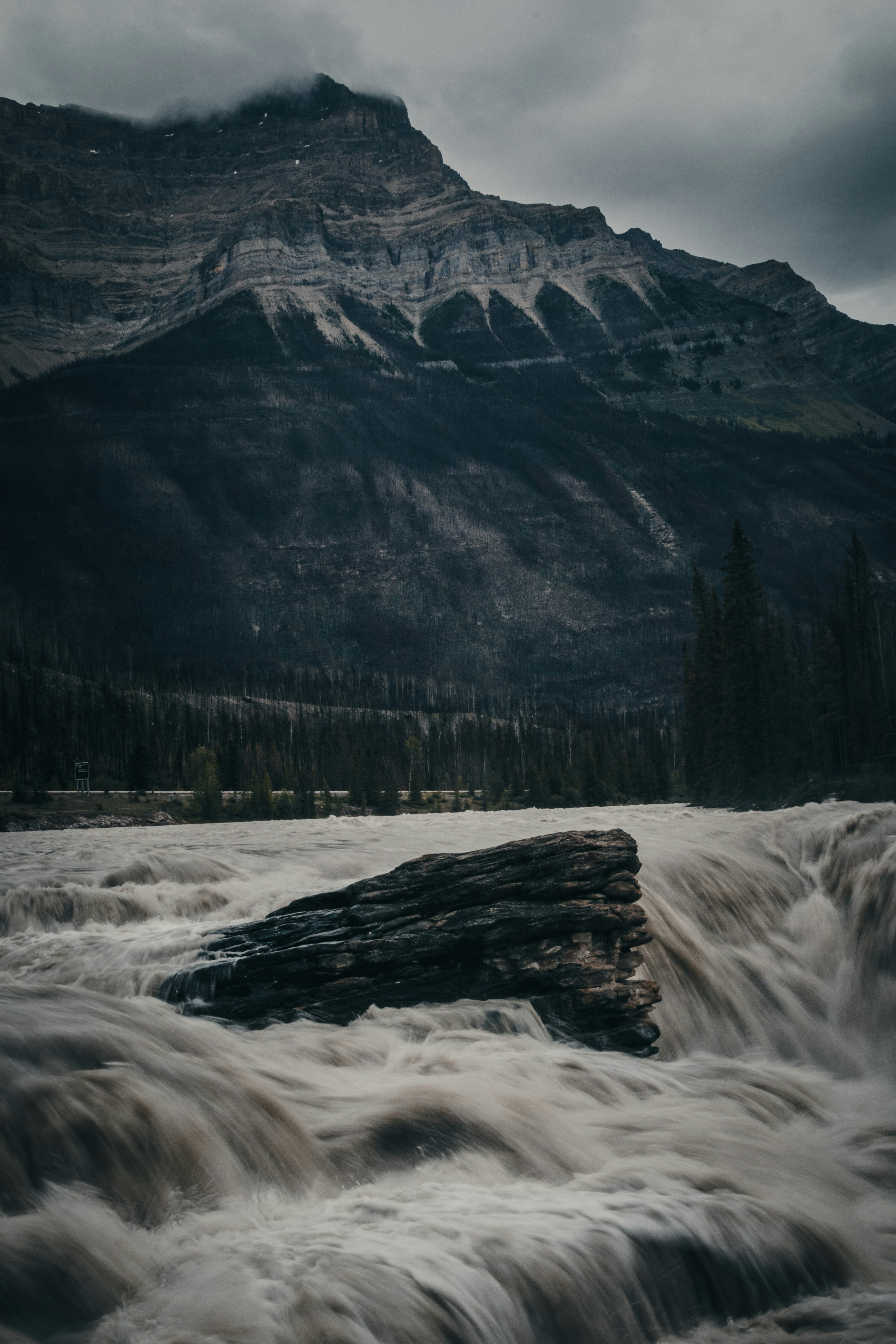 Turbulent waterfall flows past a large rock with mountains behind.