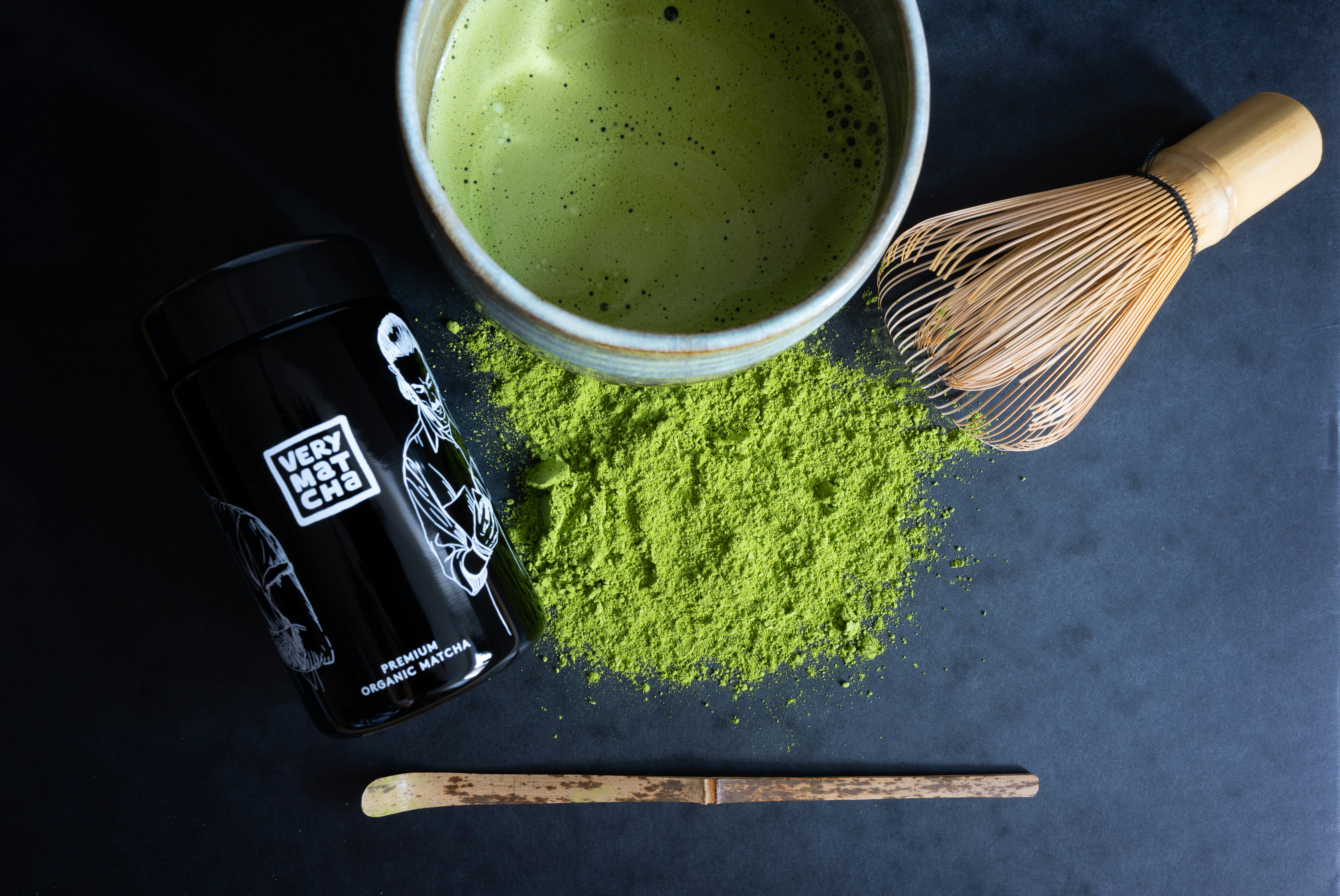 Matcha tea powder, whisk, and cup on dark surface