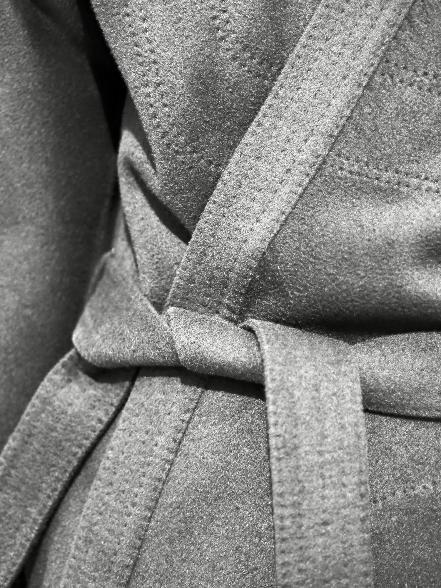 The Fellgate field coat laid flat with its belt tied, showing the stone waxed cotton and the stitched edge