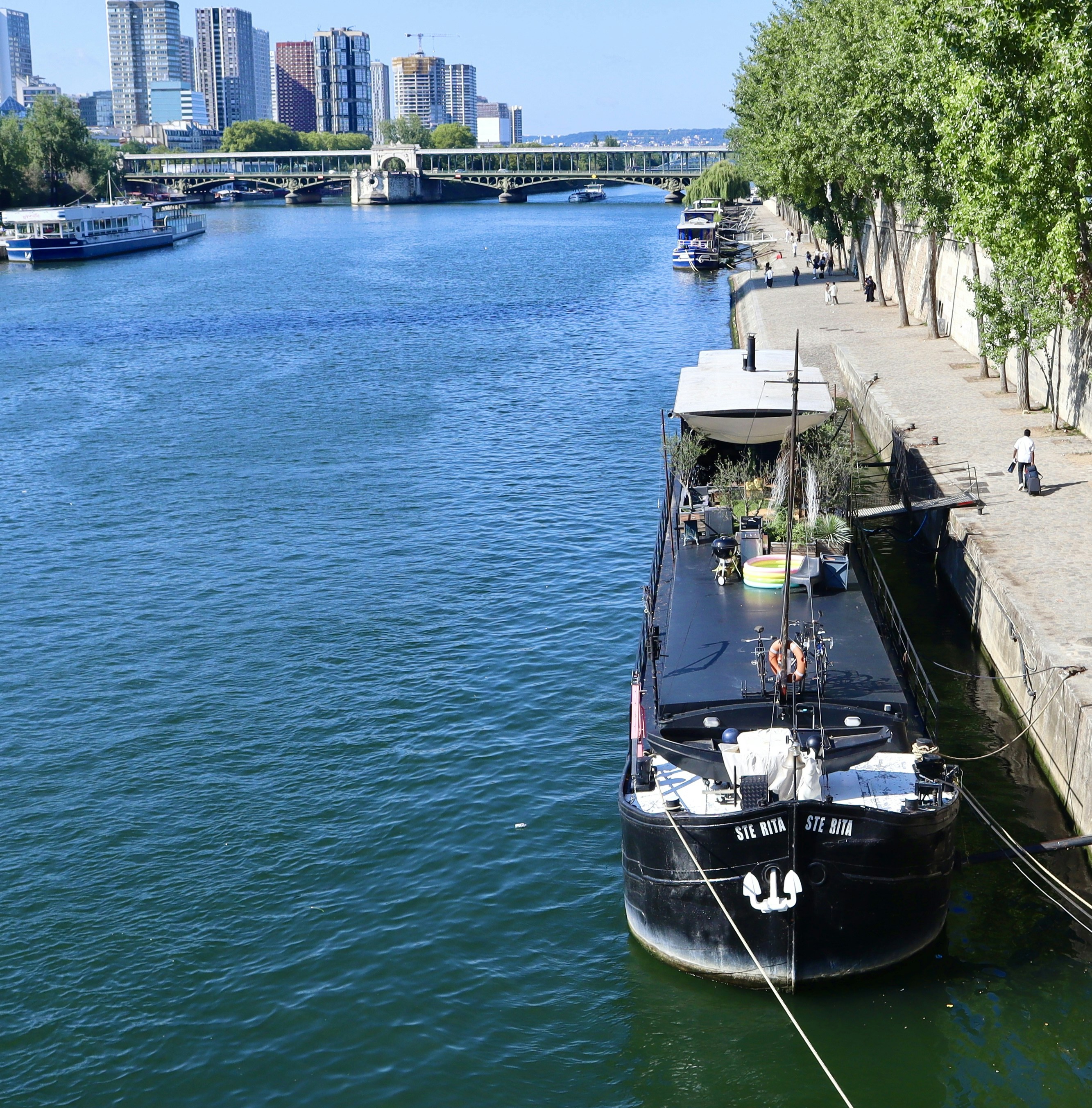 A large barge is docked along a riverbank with city buildings.
