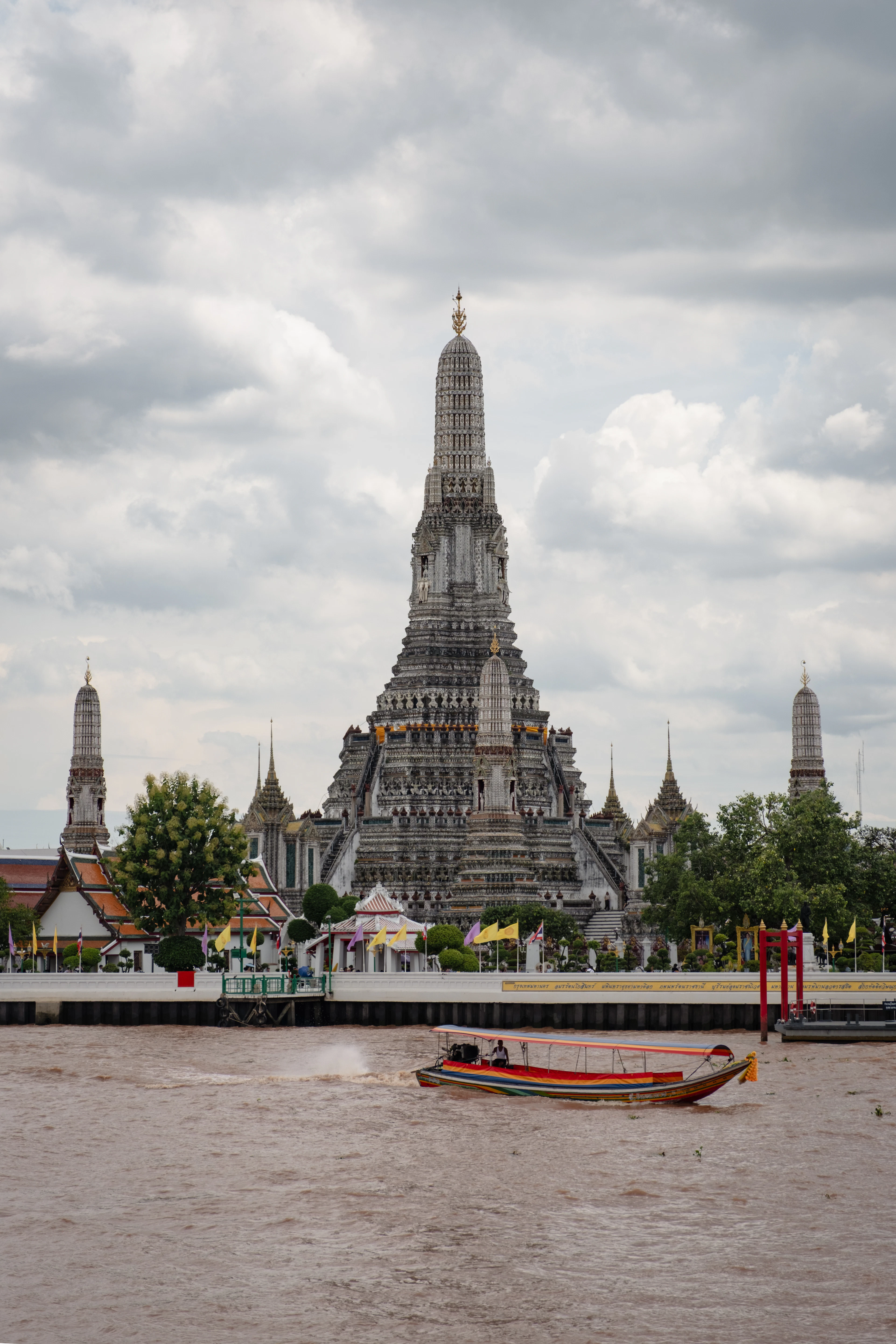 The porcelain-encrusted central prang of Wat Arun seen across the Chao Phraya River in Bangkok, with a longtail boat speeding past in the foreground