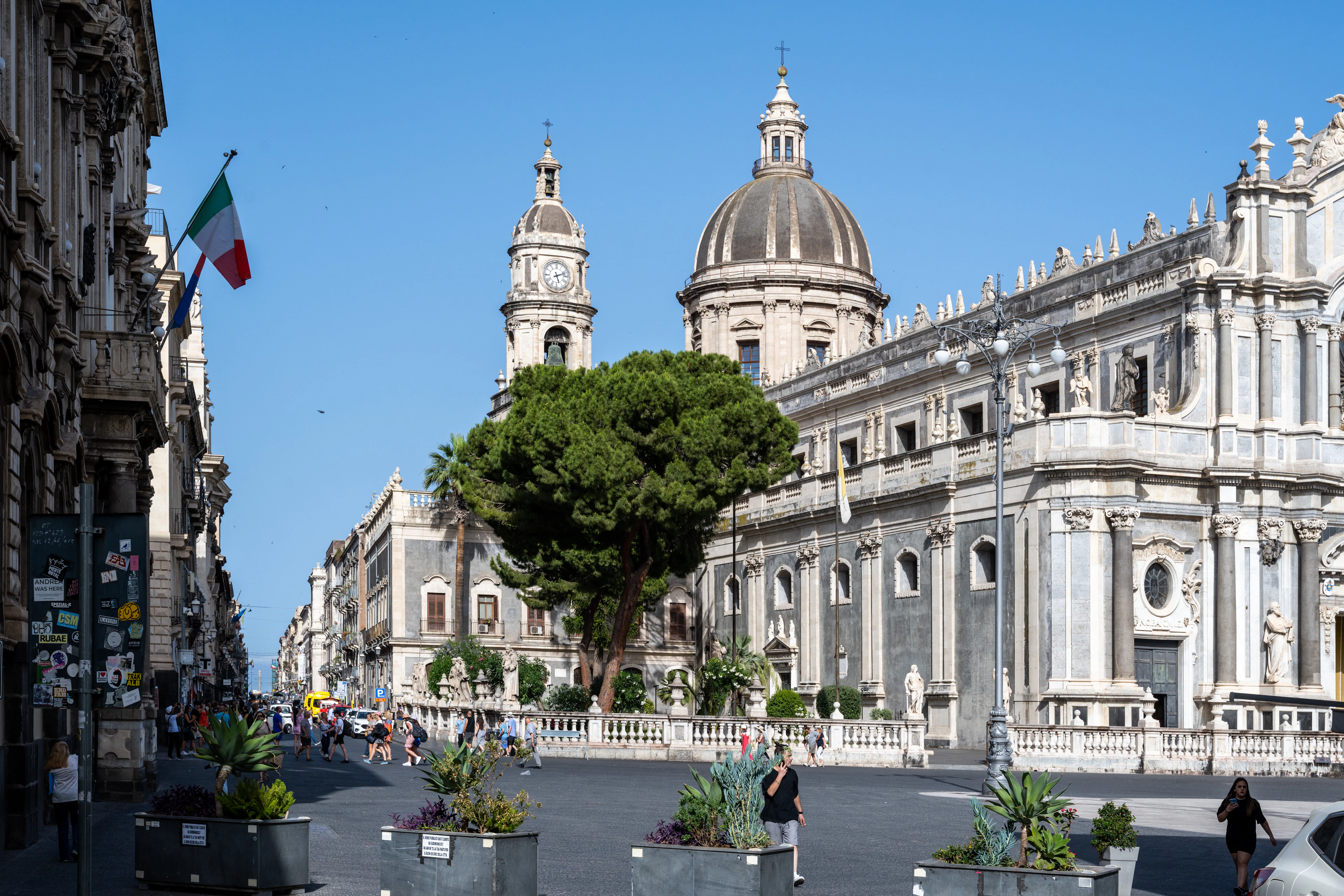 Catania's Piazza del Duomo with the cathedral, bell tower and an Italian flag under blue sky.